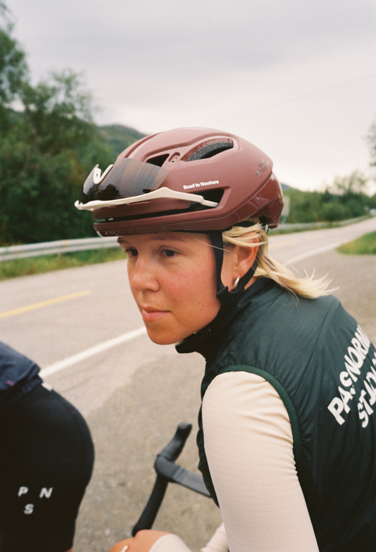 A woman wearing a maroon bicycle helmet and a black vest with 'PASSAGE' written on it, sitting on a bike on a country road surrounded by greenery.