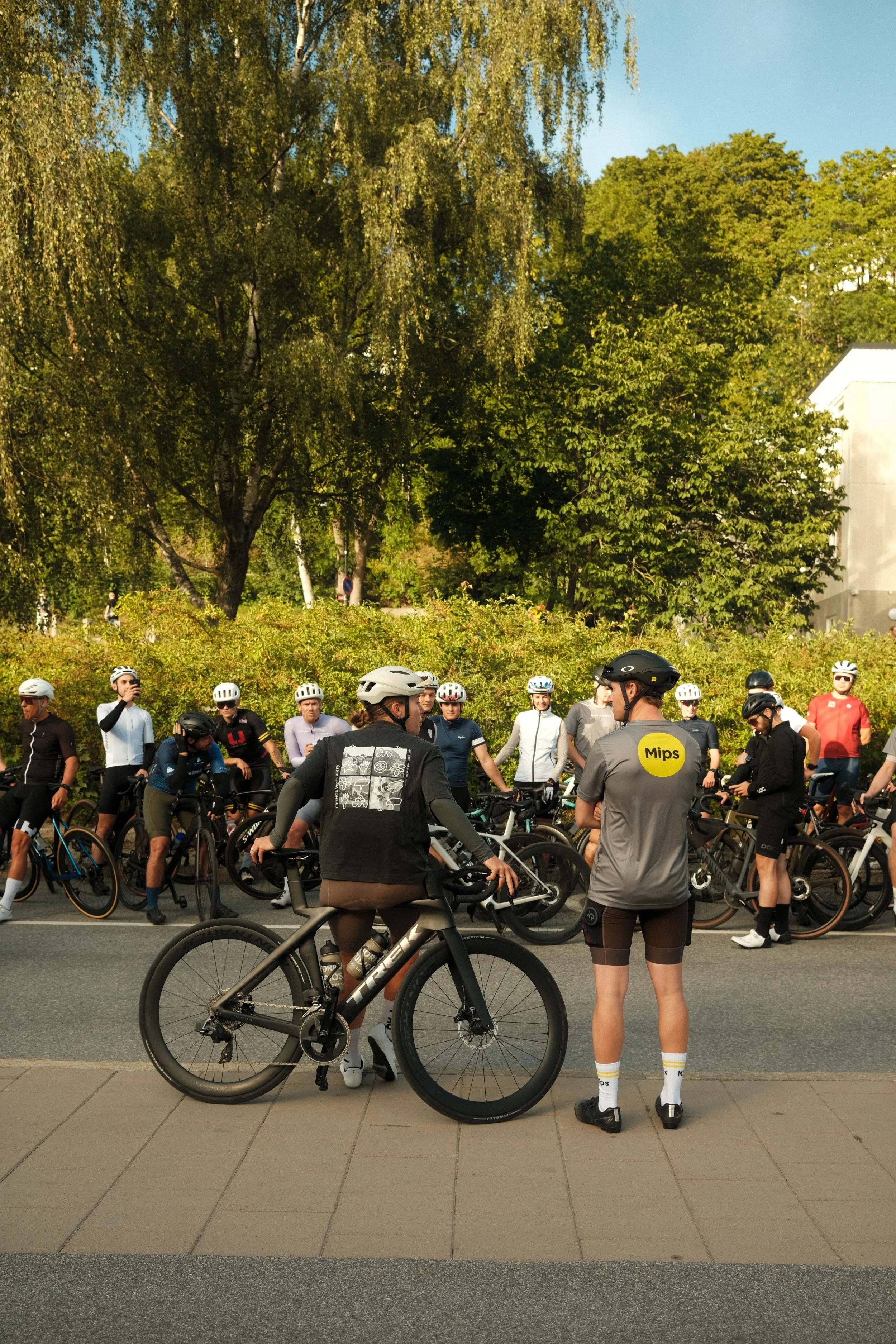 Group of cyclists wearing helmets, gathered outdoors on a sunny day, with some standing and others sitting with bikes, surrounded by green trees and a white building in the background.
