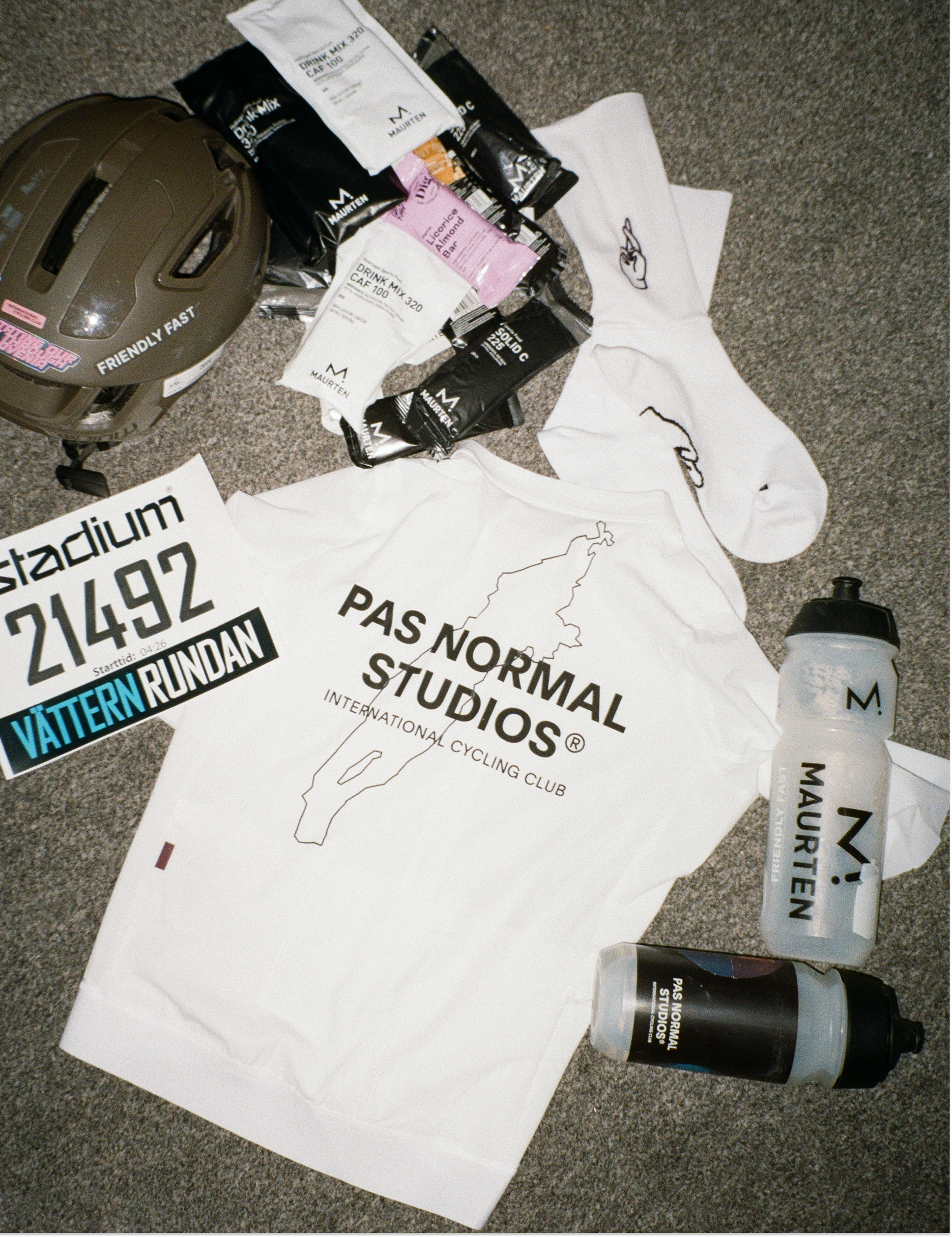 Cycling helmet, hydration bottles, cycling bibs with 'Pas Normal Studios' branding, a race bib for 'Vättern Rundan', and energy gel packets placed on a carpeted surface.