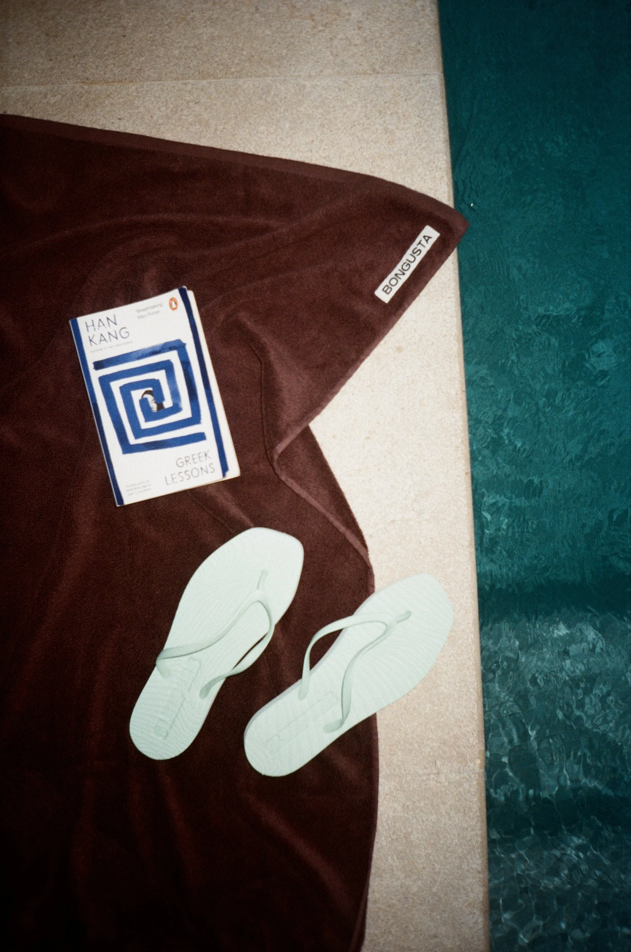 A towel, a book titled "Greek Lessons" by Han Kang, a pair of white flip-flops, and a pool are visible, suggesting a poolside setting.