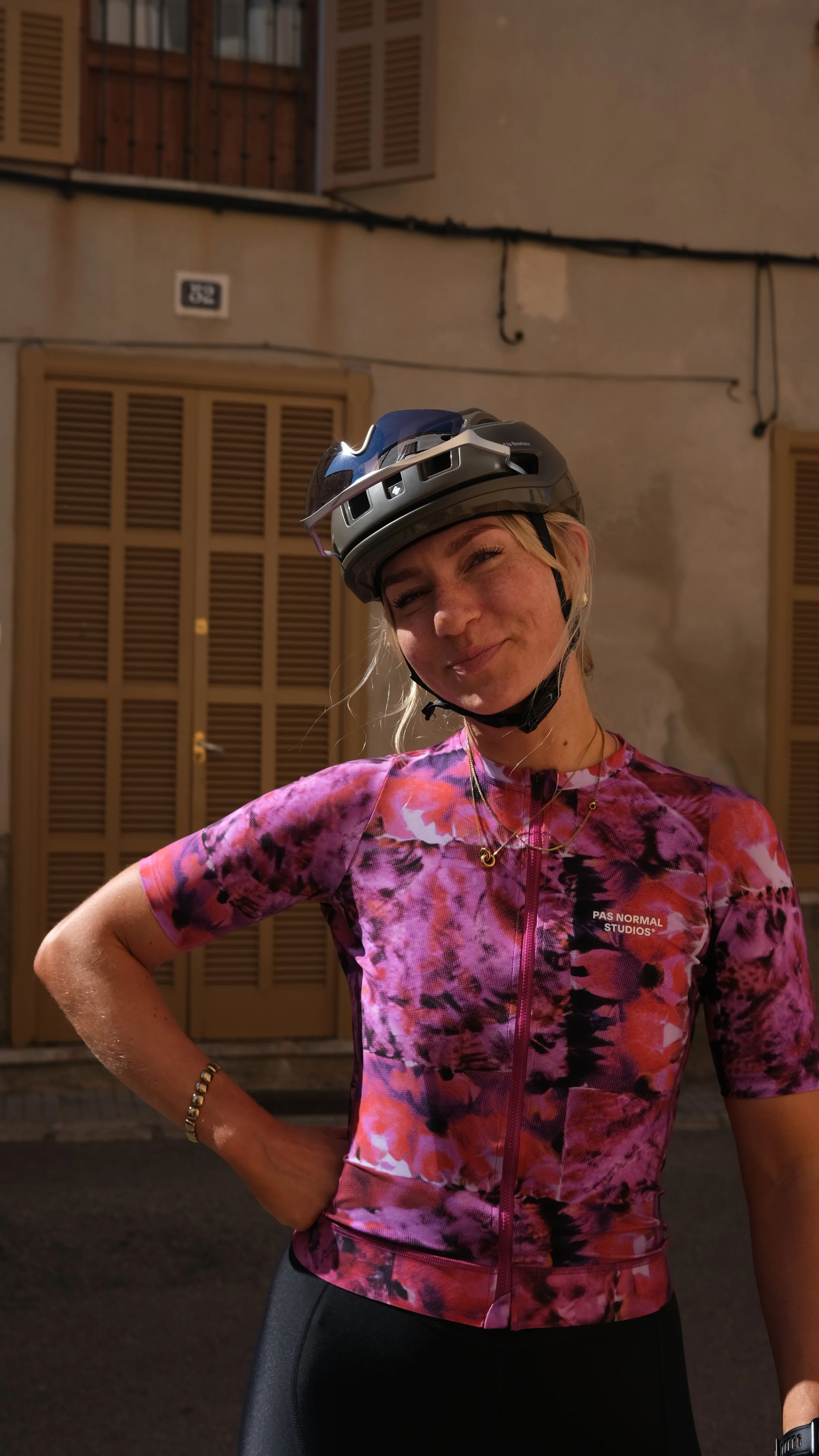 Young woman in teal cycling outfit and helmet saluting against a pink textured wall.
