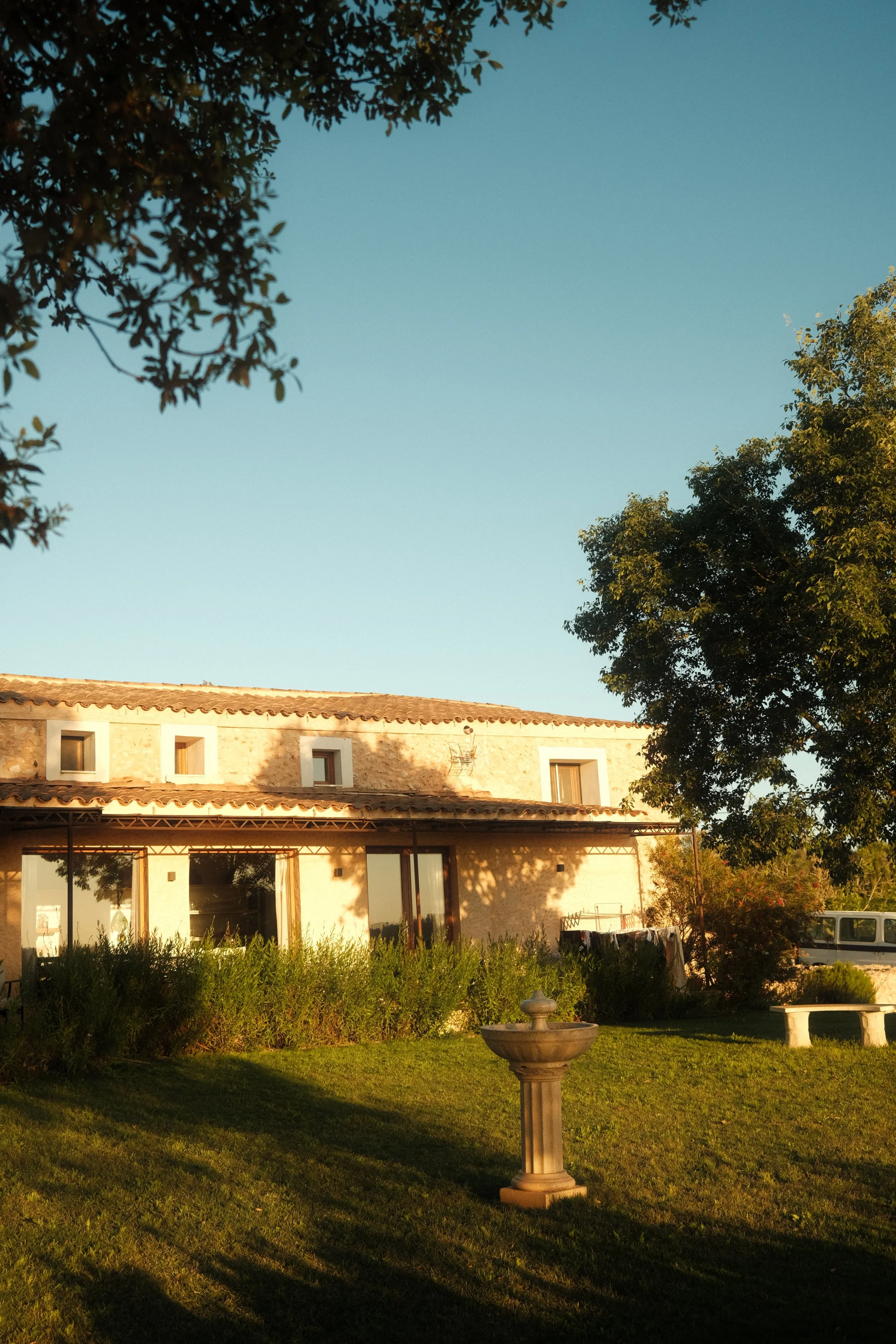 A house with a stone wall, large windows, and a tiled roof, surrounded by trees and a green lawn, with a stone birdbath and a distant vehicle in the yard, during sunset.