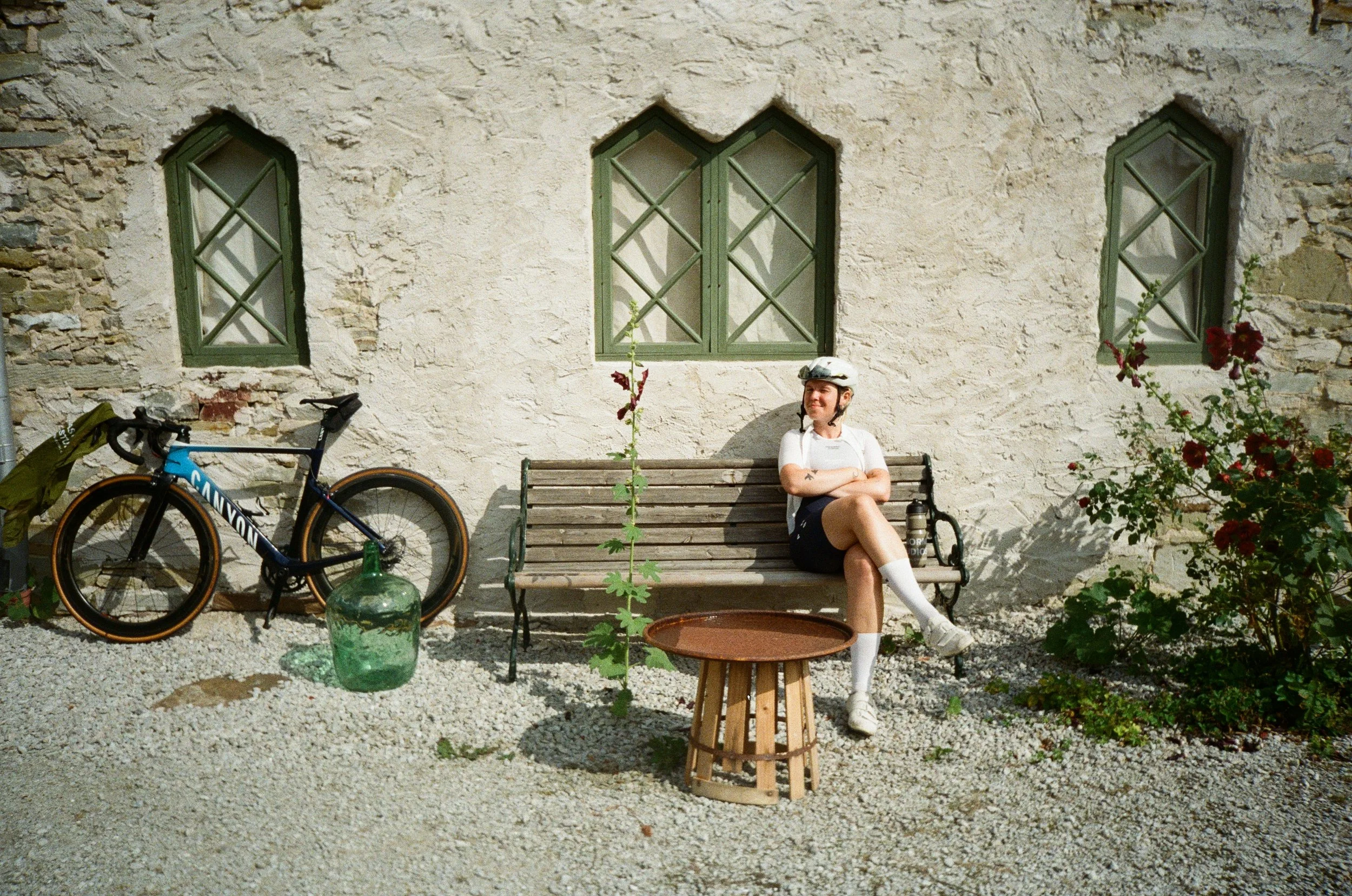 A woman in cycling gear, including a white helmet, white shirt, and black shorts, sitting on a wooden bench outside a stone building with three green-framed windows. Her arms are crossed, and she is smiling. A blue bicycle with a green bag attached to it leans against the building, and there is a large green glass jar, a wooden structure, and a tall plant growing from a pot nearby. Red flowers are on the right side of the image.