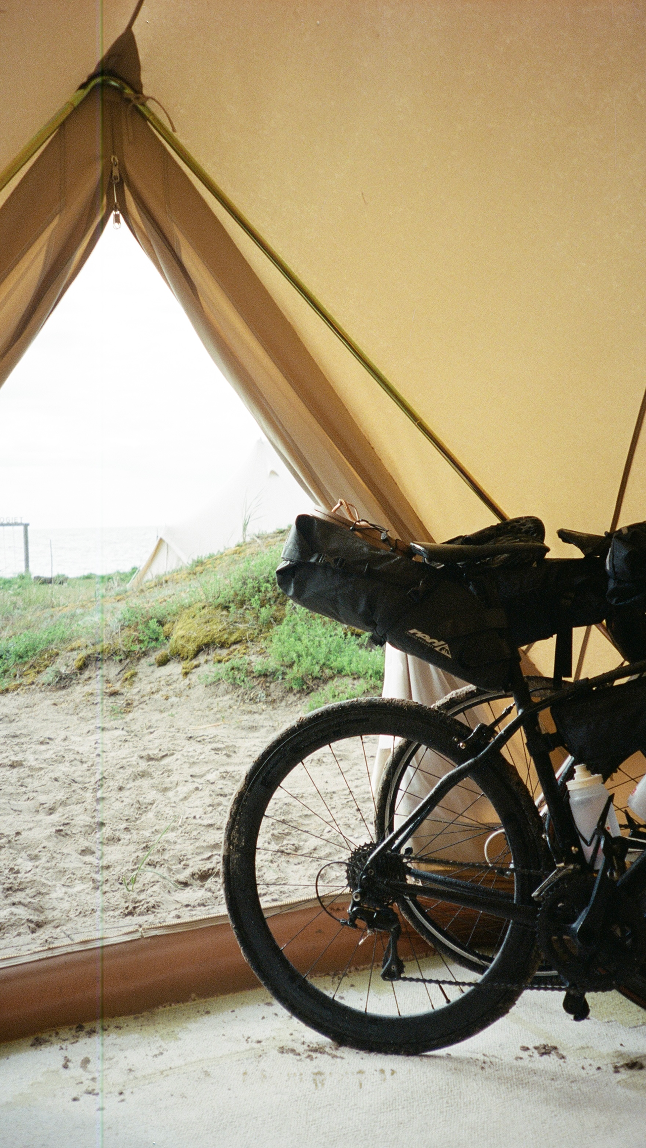 Interior of a beach tent with a bicycle inside, facing a sandy area with green grass and a fence near the ocean.