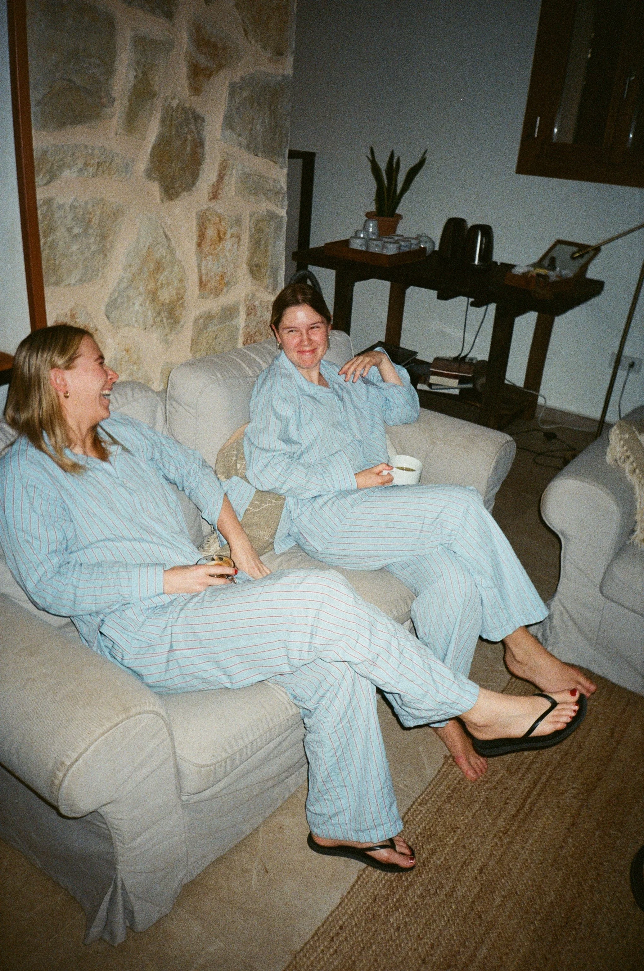 Two women in striped pajamas sitting on a sofa, laughing, one holding a TV remote and the other holding a cup, in a cozy living room with a stone wall background.