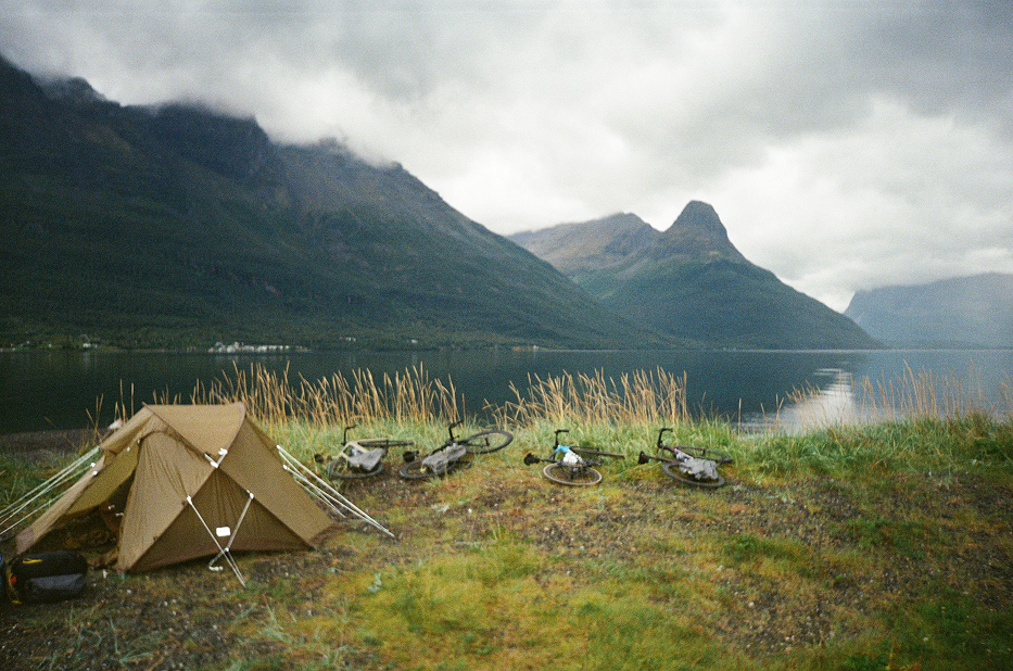 A camping scene by a lake with a tent and three bicycles on a grassy area, mountains in the background, and overcast skies.