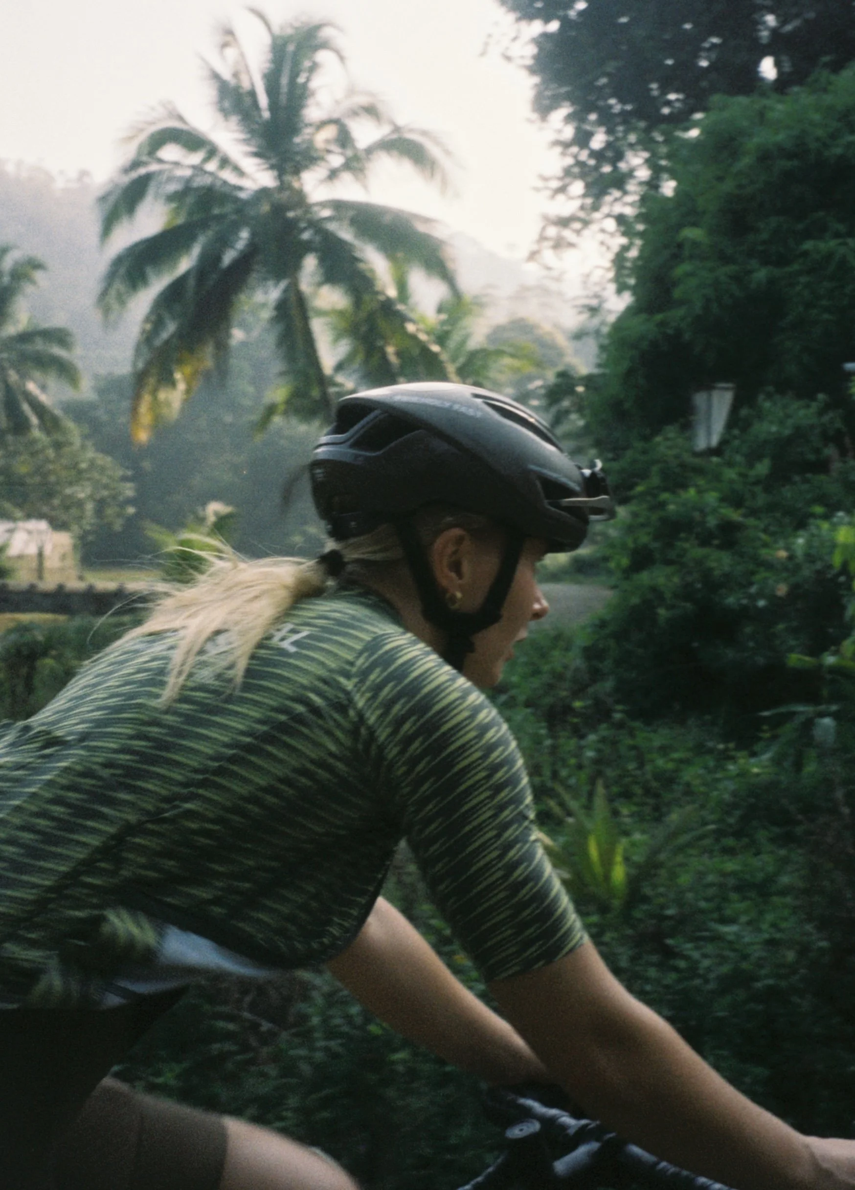 A woman wearing a black bicycle helmet and a green and black striped shirt riding a bicycle through a lush, tropical landscape with palm trees and dense greenery.