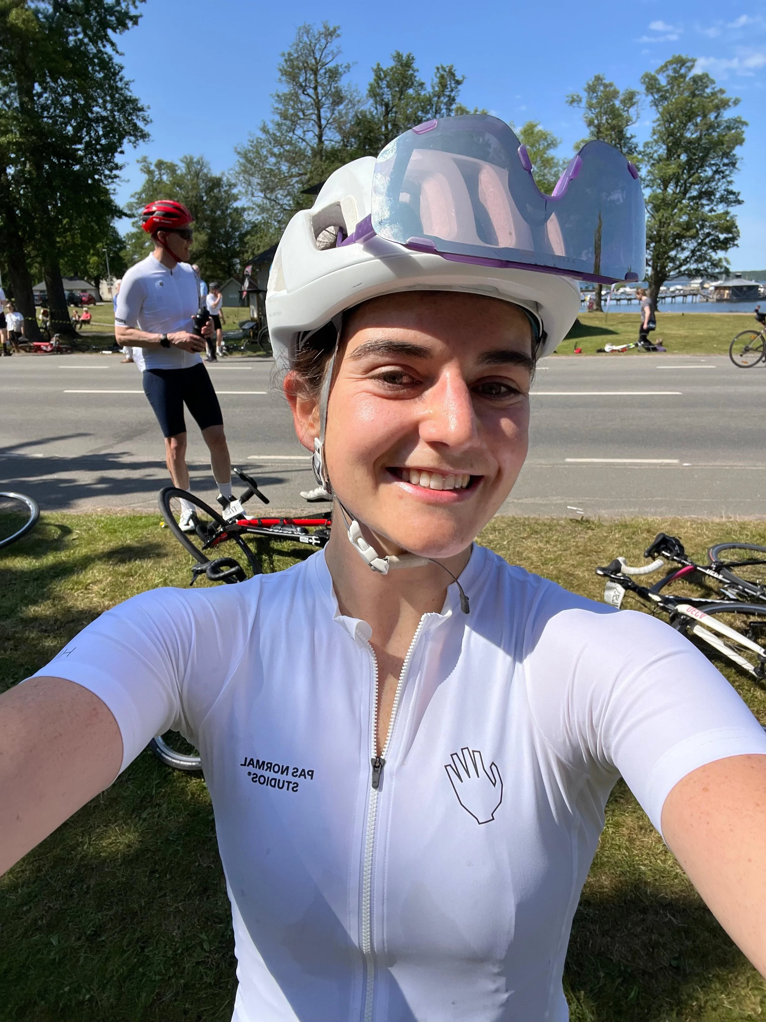 A smiling woman in a white cycling jersey and helmet taking a selfie outdoors on a sunny day, with bicycle gear and other cyclists in the background near a grassy area and a road