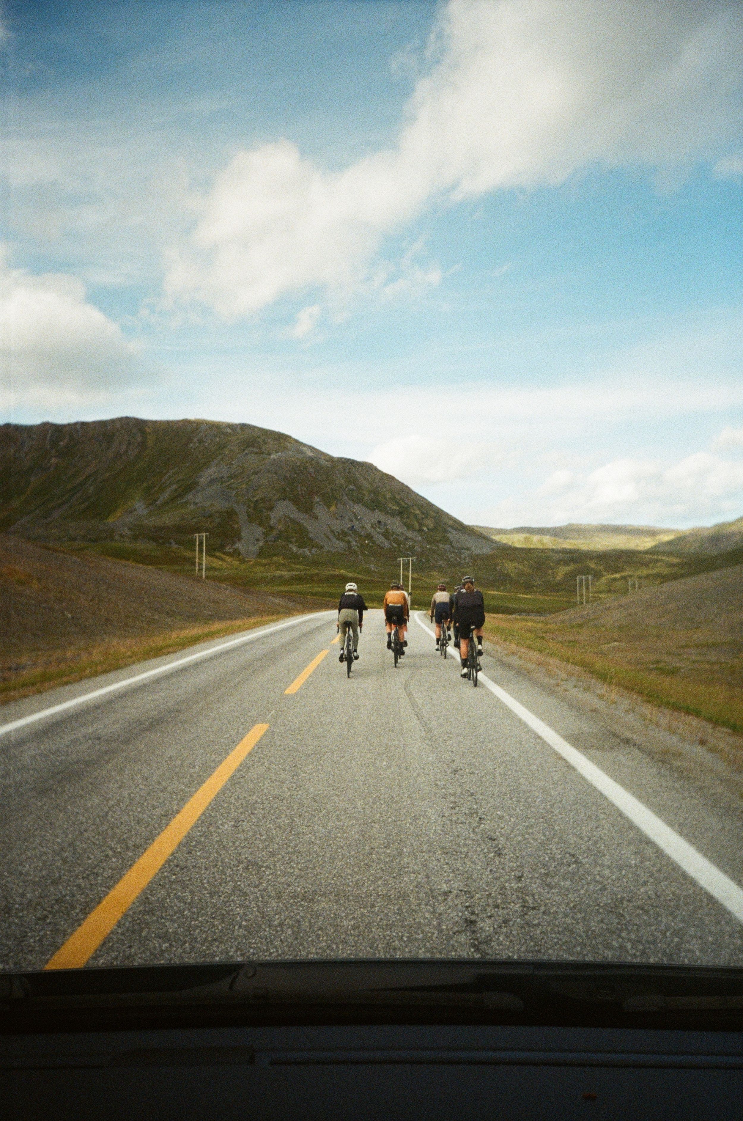 Four cyclists riding on a rural mountain road under a cloudy sky with hills in the background.