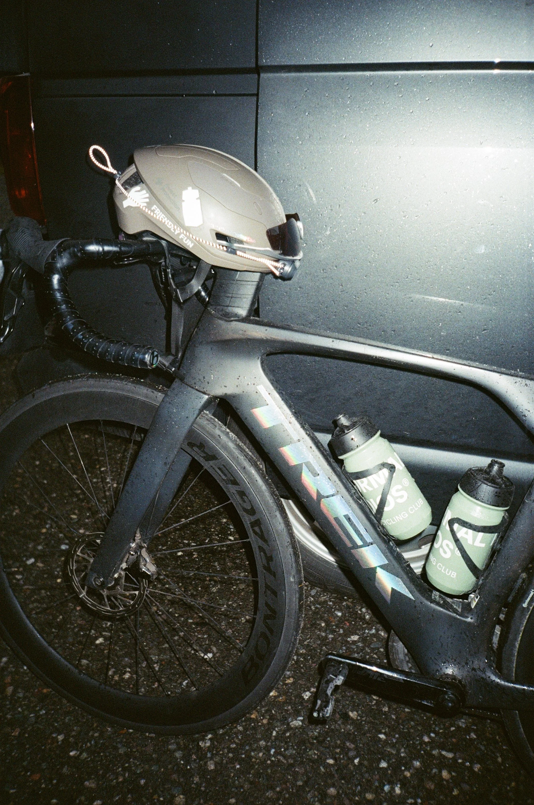 A gray bike with two water bottles attached to the frame, a helmet hanging on the handlebar, and a black emblem on the front fork, parked in front of a dark gray vehicle.