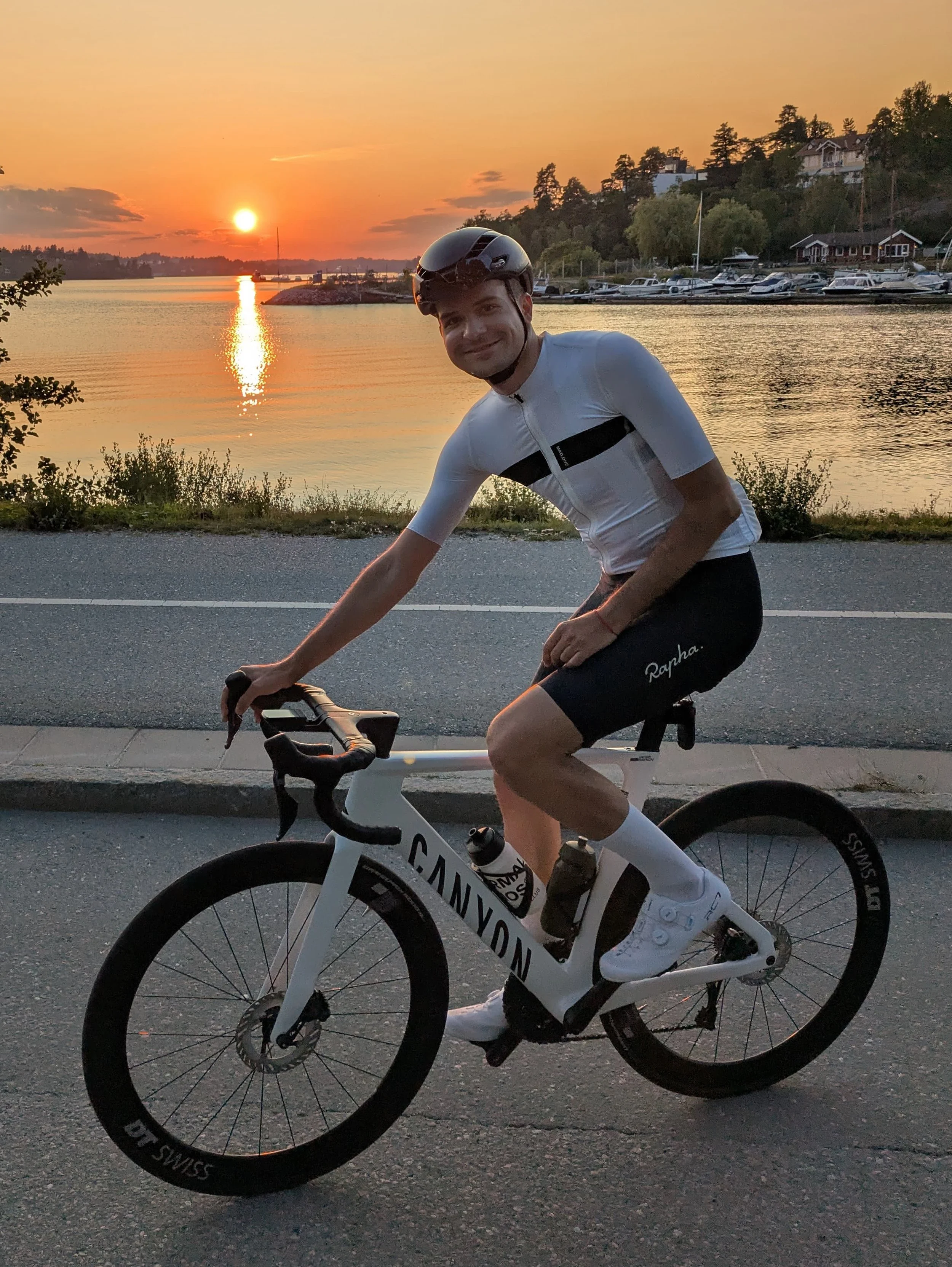 A man wearing a white cycling outfit and a black helmet riding a white Canyon road bike along a paved road near a body of water at sunset.