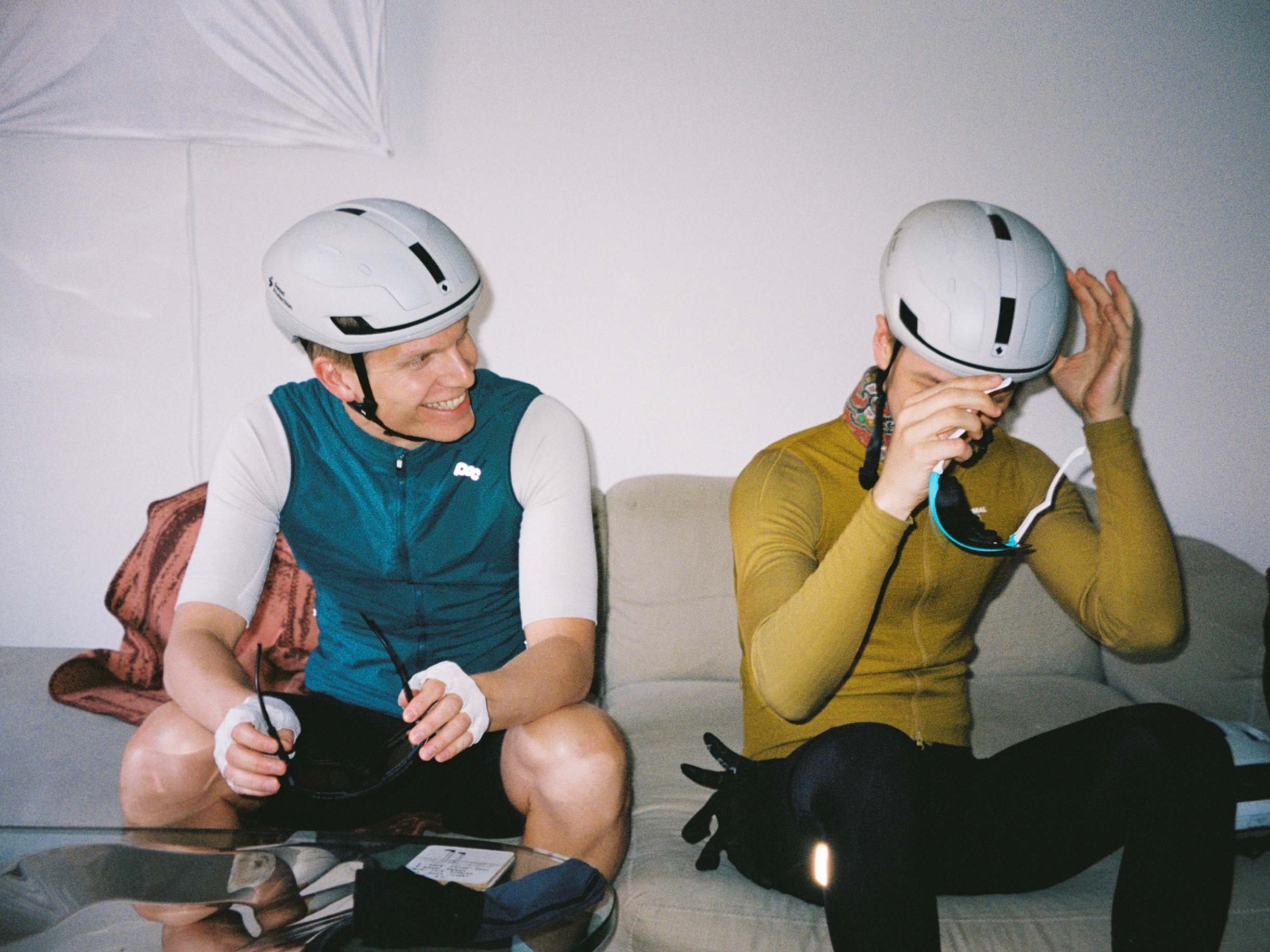 Two people sitting on a couch, wearing white bicycle helmets. The person on the left is smiling and holding sunglasses, wearing a blue and white cycling outfit. The person on the right is adjusting their helmet, wearing a yellow long-sleeve shirt and black pants.