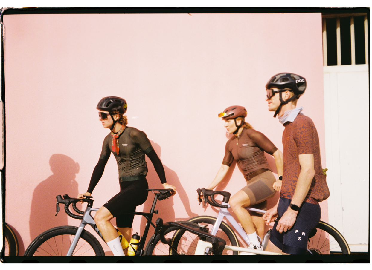 Three cyclists in helmets and cycling gear riding stationary bikes indoors against a pink background.