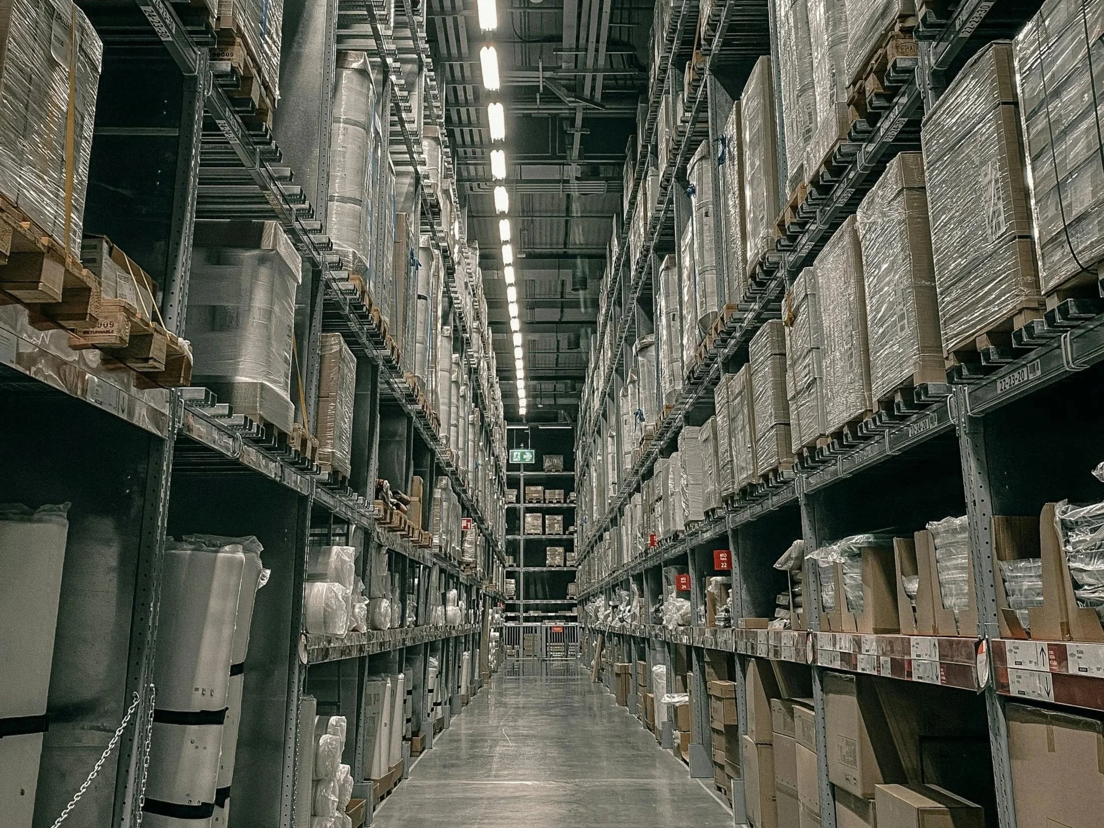 Aisle in a warehouse with tall shelves stocked with boxed products, seen from a central walkway under bright overhead lighting.