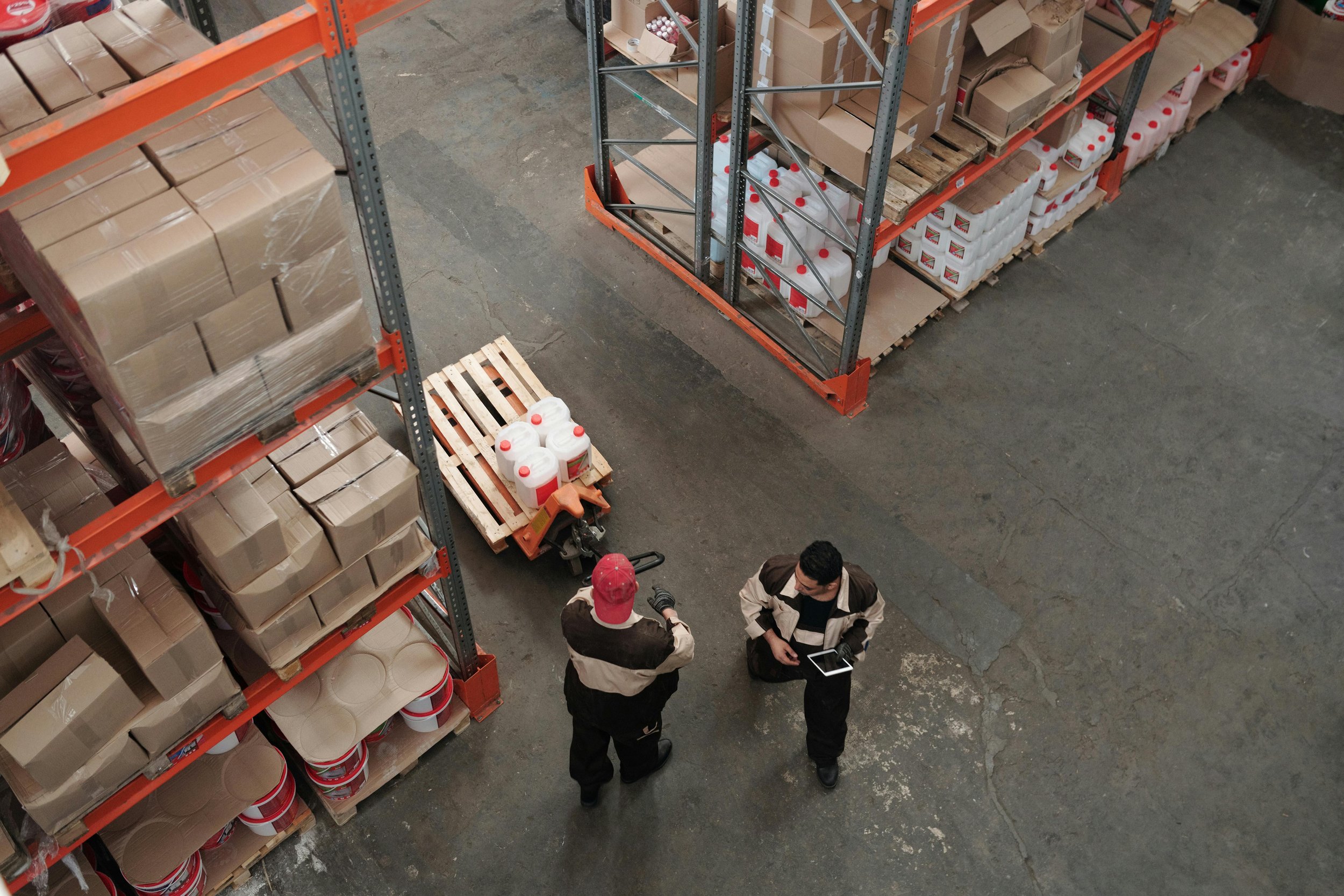Two warehouse workers in uniform conversing in an industrial storage area with shelves of boxes, some on pallets and a pallet of gallon containers nearby.