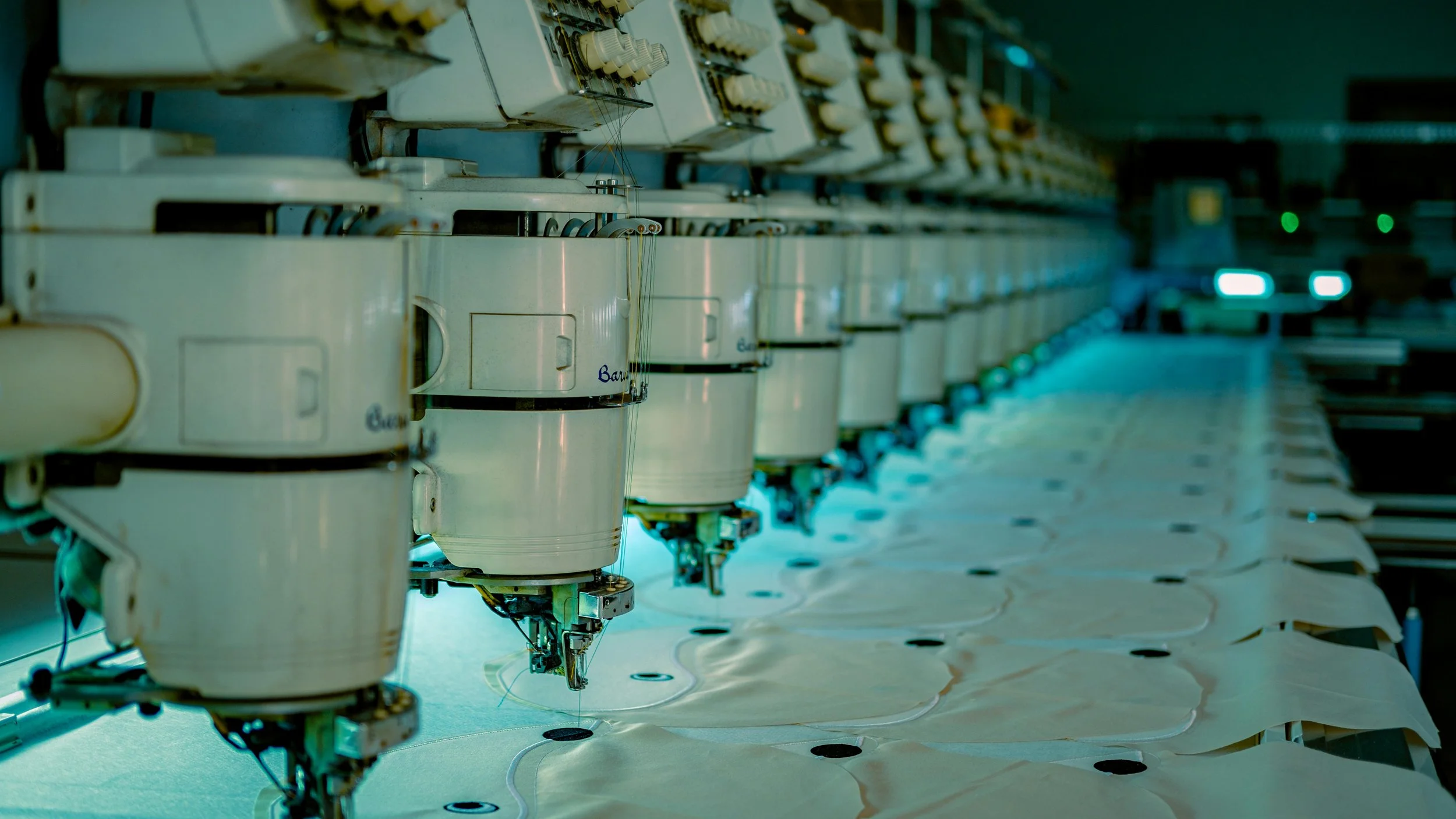 Close-up of multiple industrial sewing machines with fabric feeding through them, in a factory setting with a vehicle in the background.