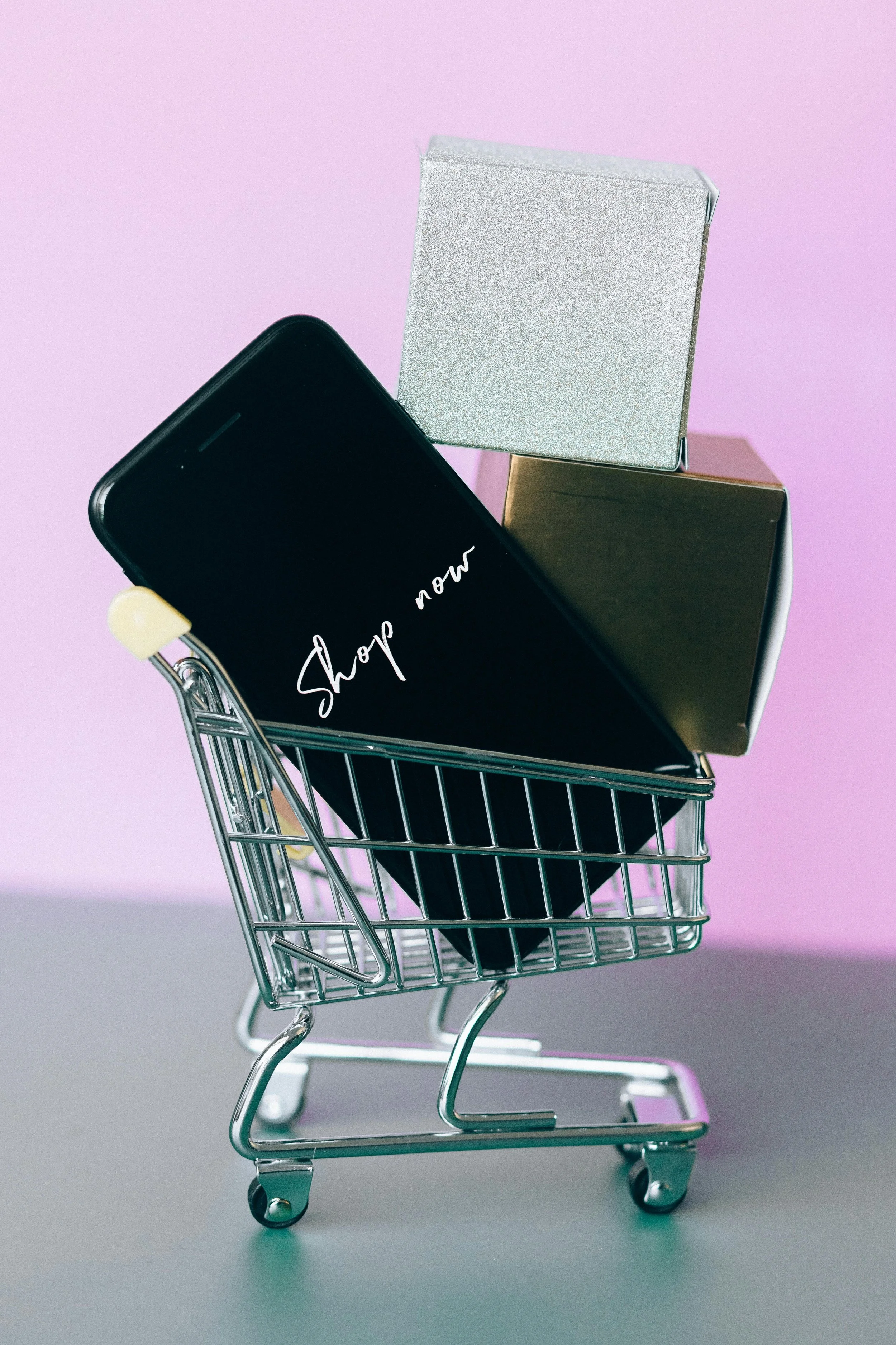 Miniature shopping cart with a smartphone displaying 'Shop now' and two small gift boxes, on a pink background.