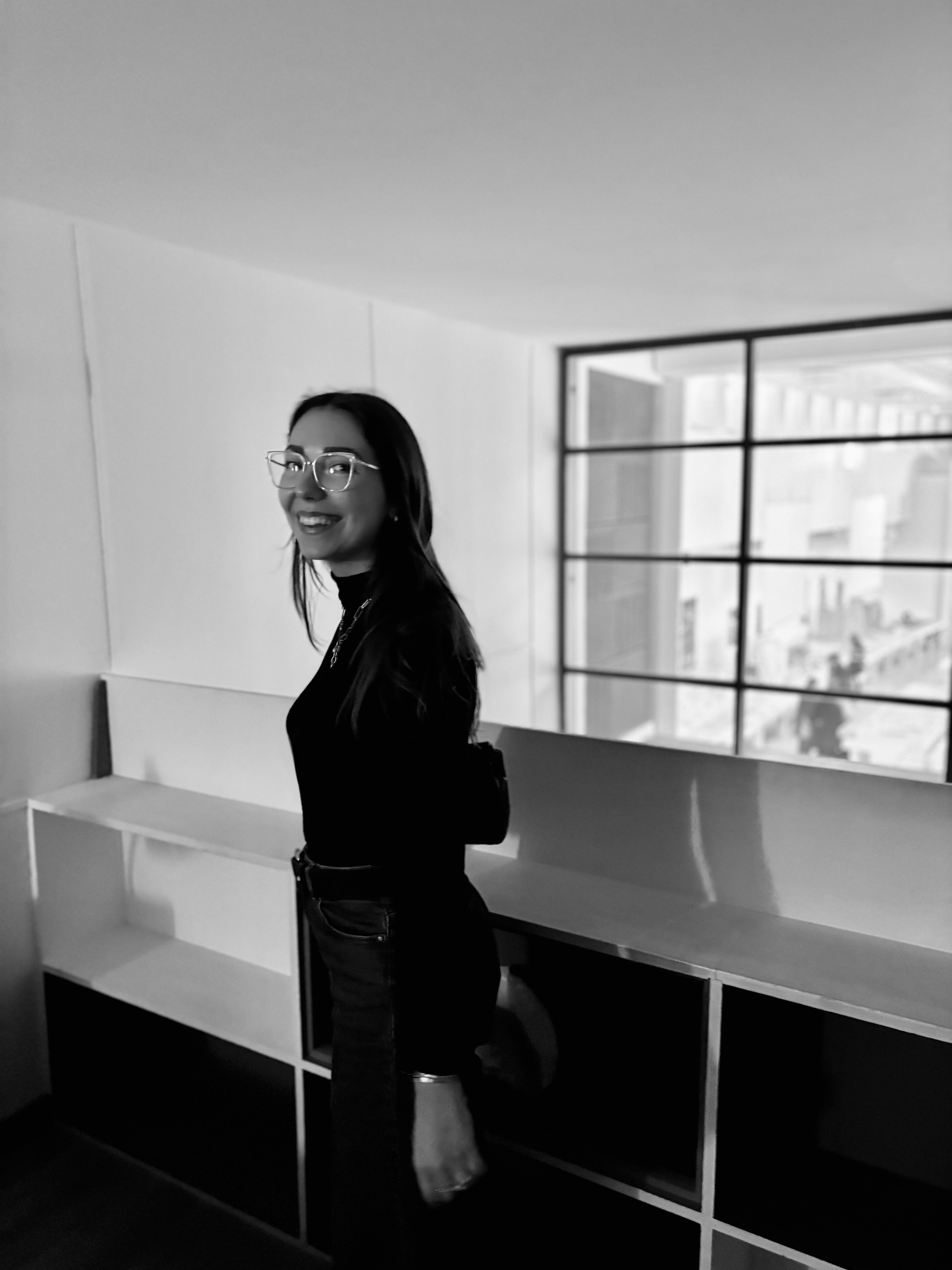 Une femme portant des lunettes, vêtue d'une veste noire, se tient dans une pièce moderne avec un grand mur de fenêtres laissant entrer beaucoup de lumière naturelle. Elle sourit et fait face à la caméra.