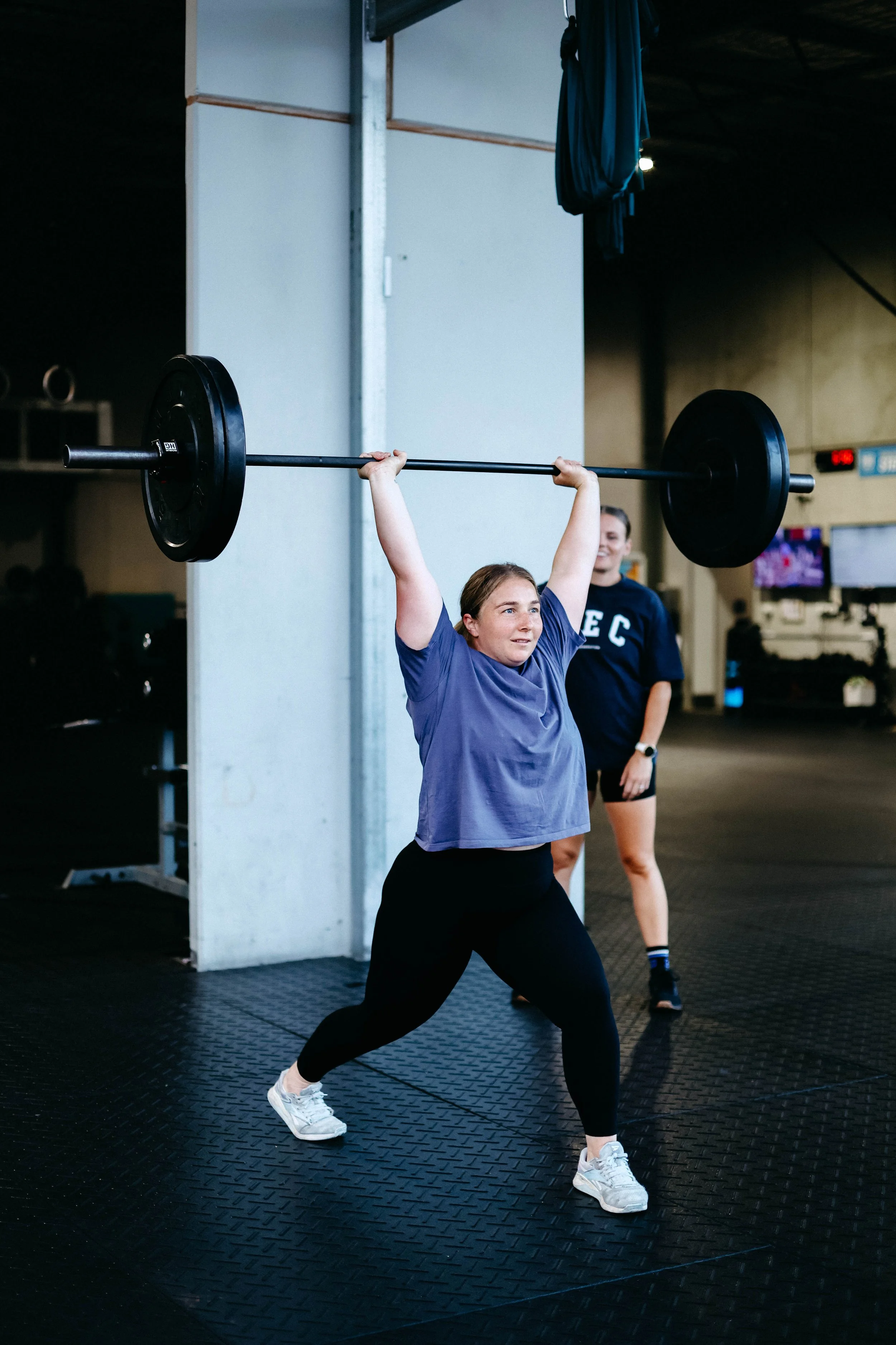 A woman lifting a barbell overhead at a gym, with a person in the background smiling.