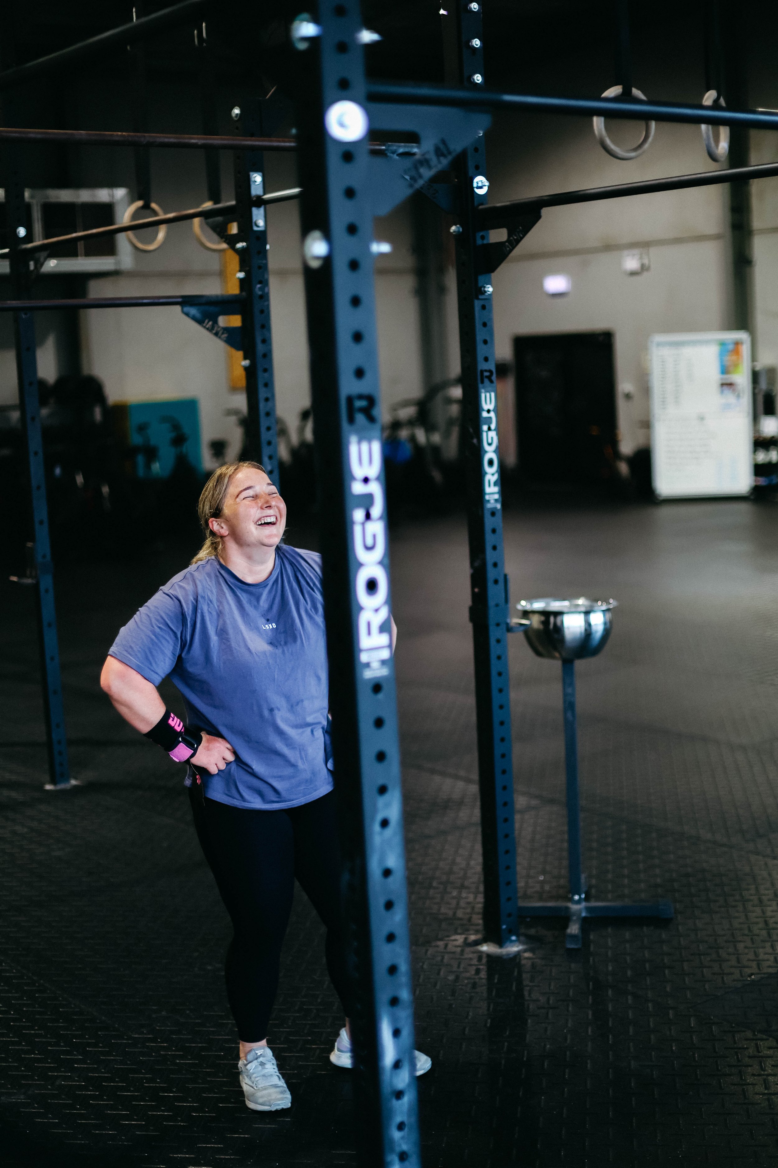 A woman laughing inside a gym, standing near a blue Rogue-branded pull-up rig with gymnastic rings hanging from it.
