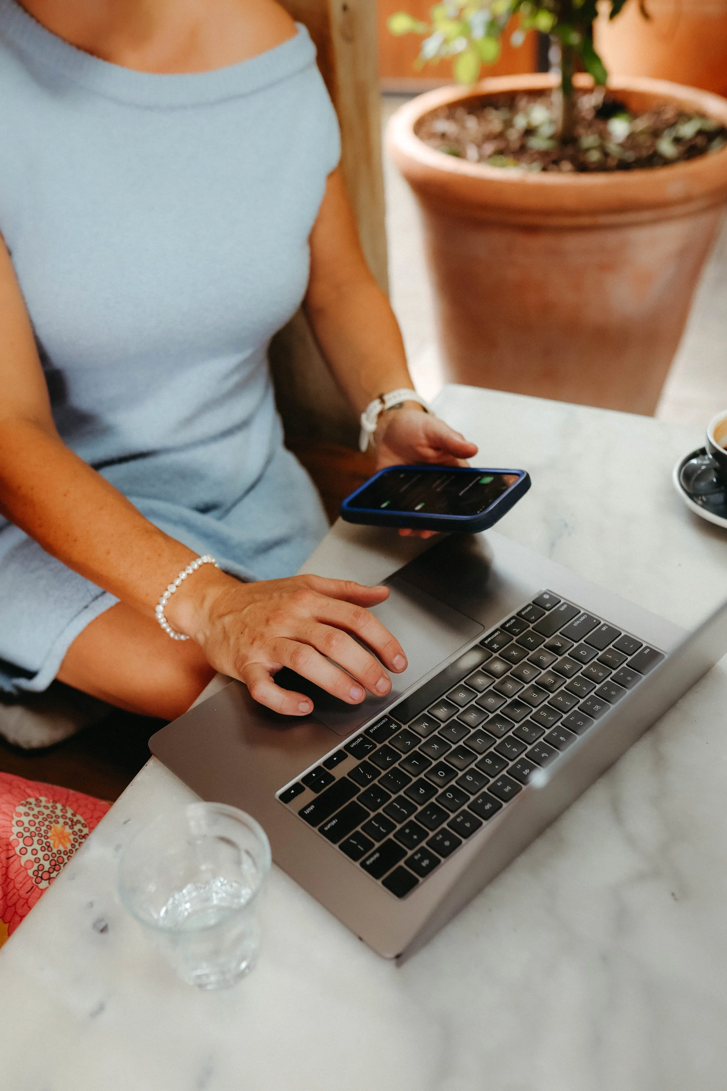Katelyn sitting at a table at local Dubbo Cafe, Press, with a laptop, holding a smartphone. There is a glass of water, a cup of coffee, and a potted plant in the background.