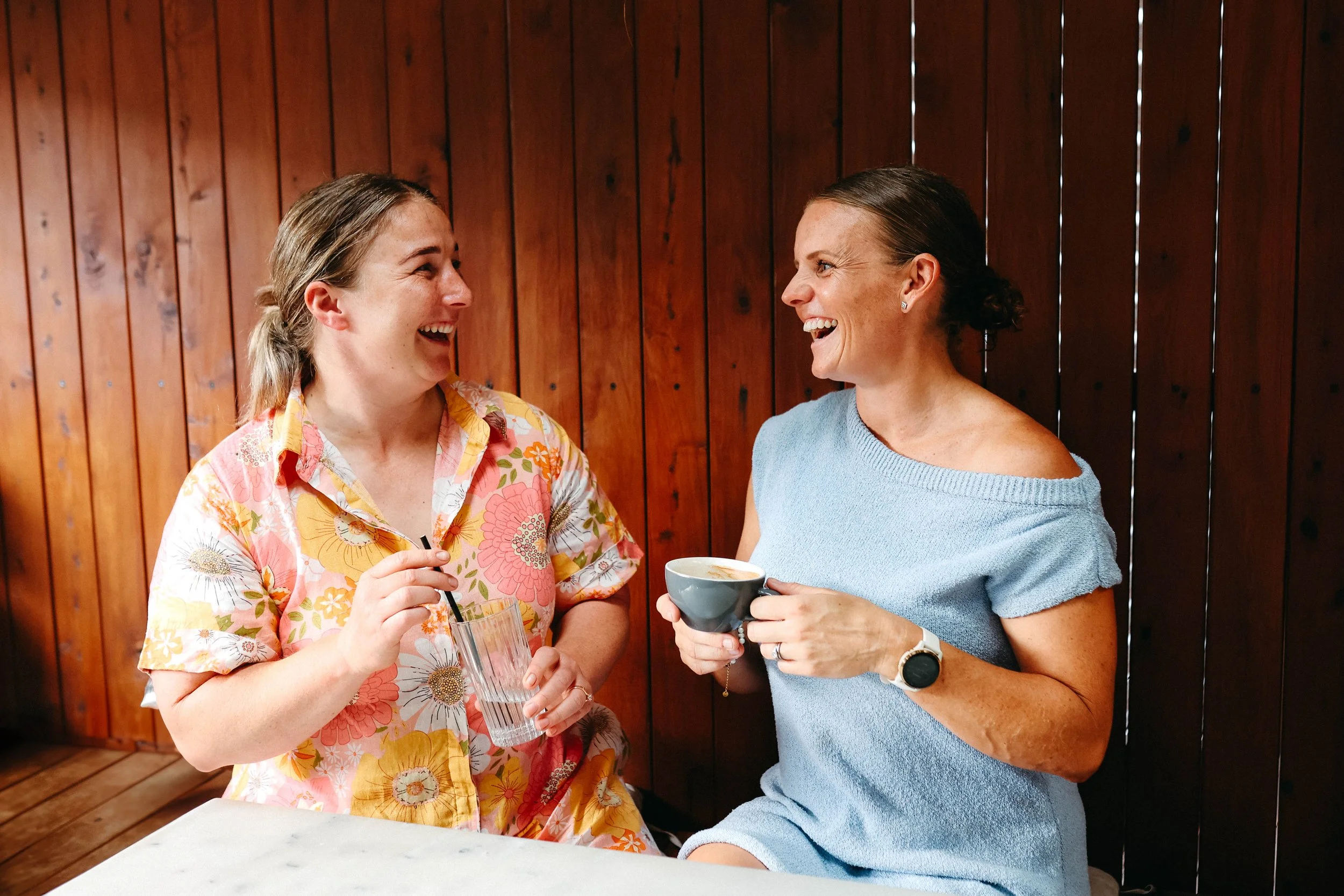Two women are sitting at a table, smiling and laughing while talking. One woman is holding a glass of water, and the other is holding a mug. They are in a room with wooden walls.