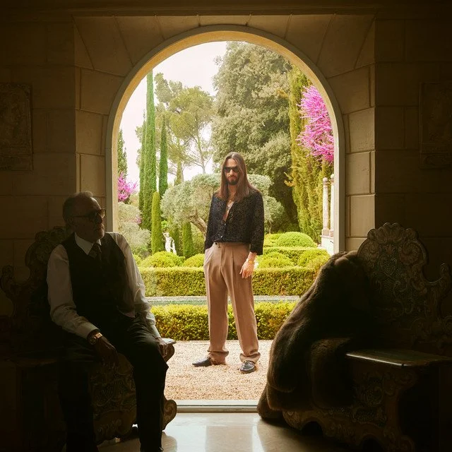 Trois personnes dans un cadre extérieur visible à travers une grande arche en pierre. Deux hommes assis à l'intérieur, un troisième homme se tenant derrière la porte, dans un jardin avec des arbres et des fleurs colorées.