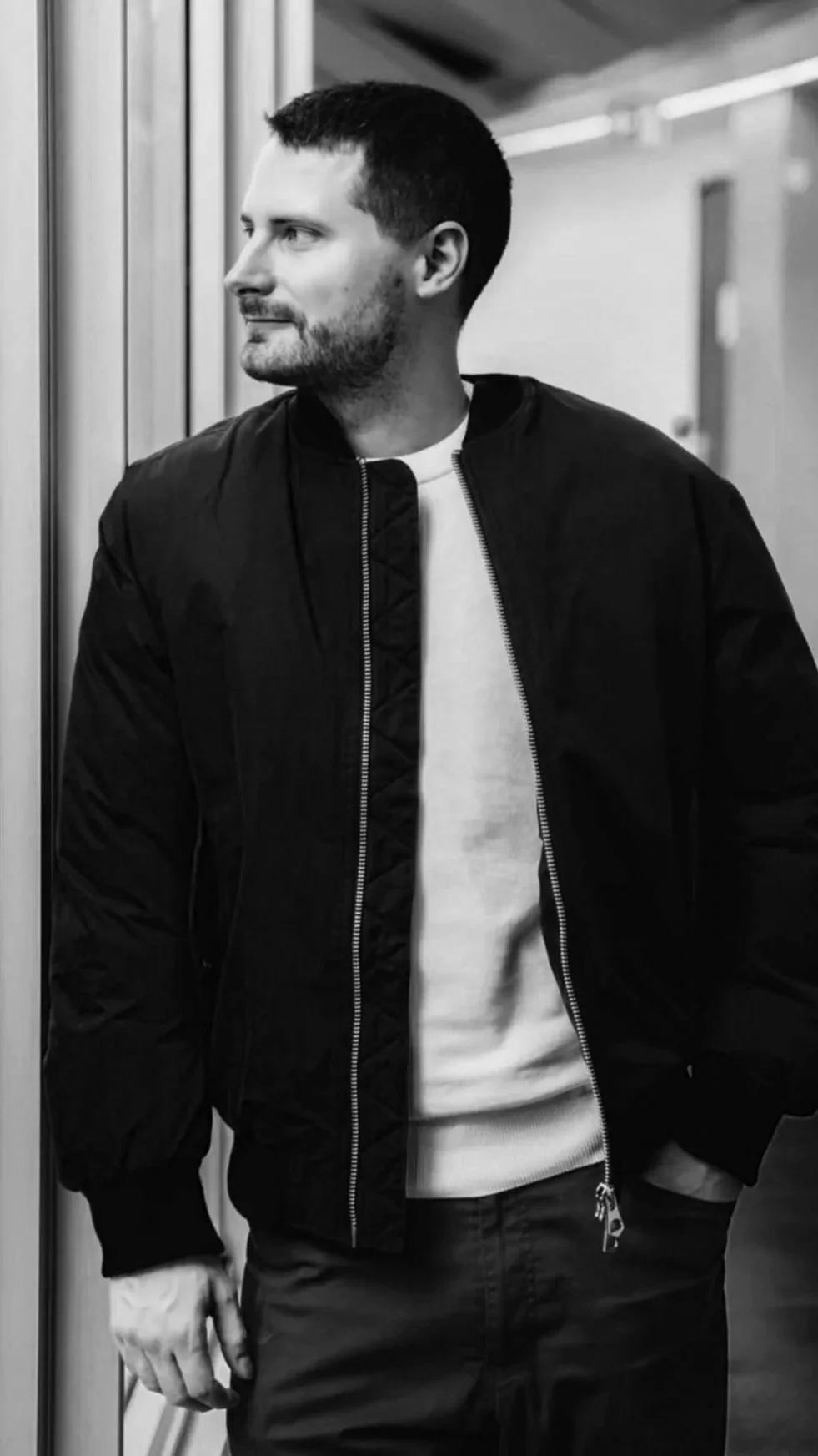 Un homme avec une barbe, portant une veste noire et un t-shirt blanc, regarde vers la gauche dans une ambiance monochrome.