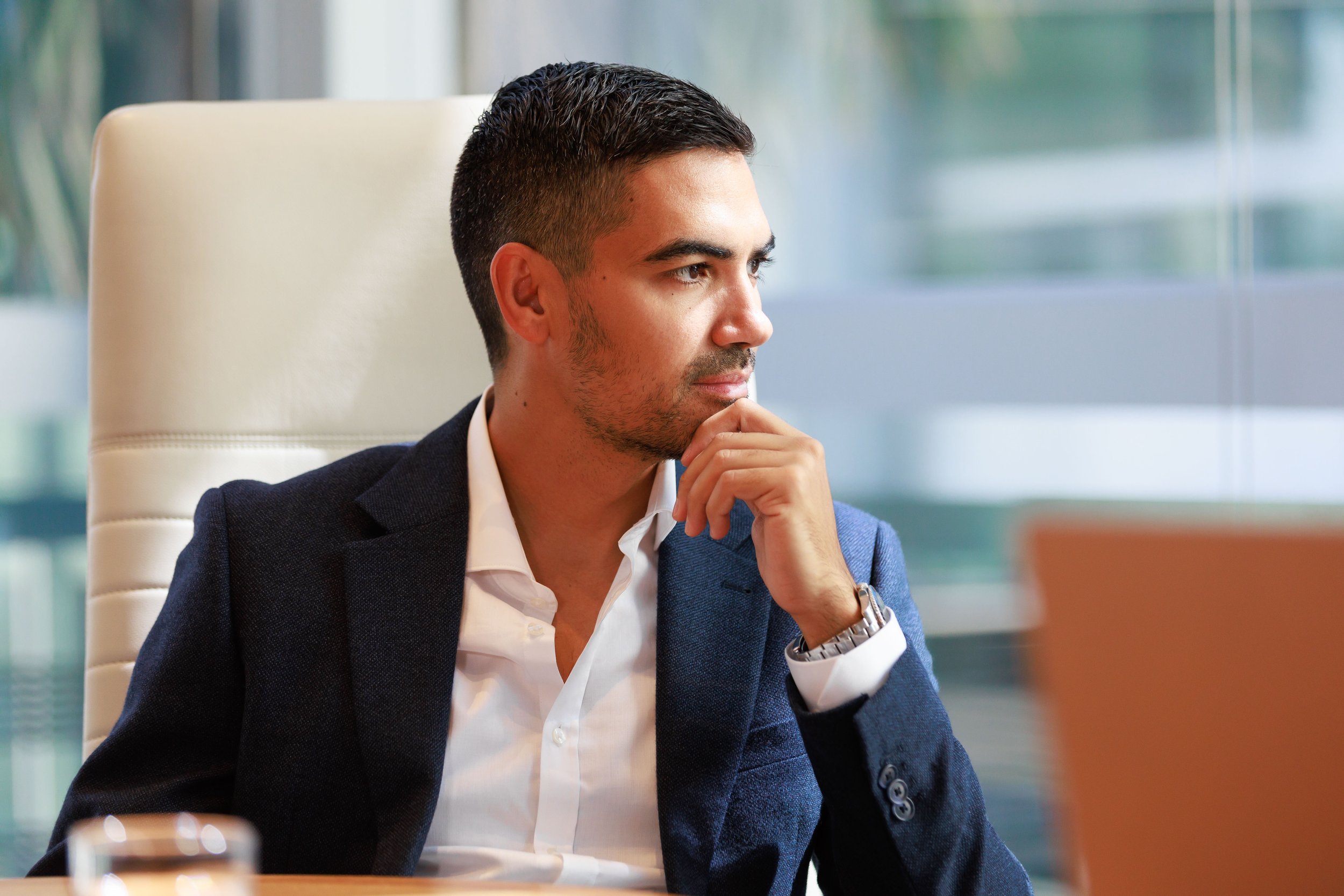 A man with dark hair, a beard, and light skin is sitting at a desk in a modern office, wearing a navy blazer and white shirt, with a thoughtful expression, resting his chin on his hand and looking to the side.