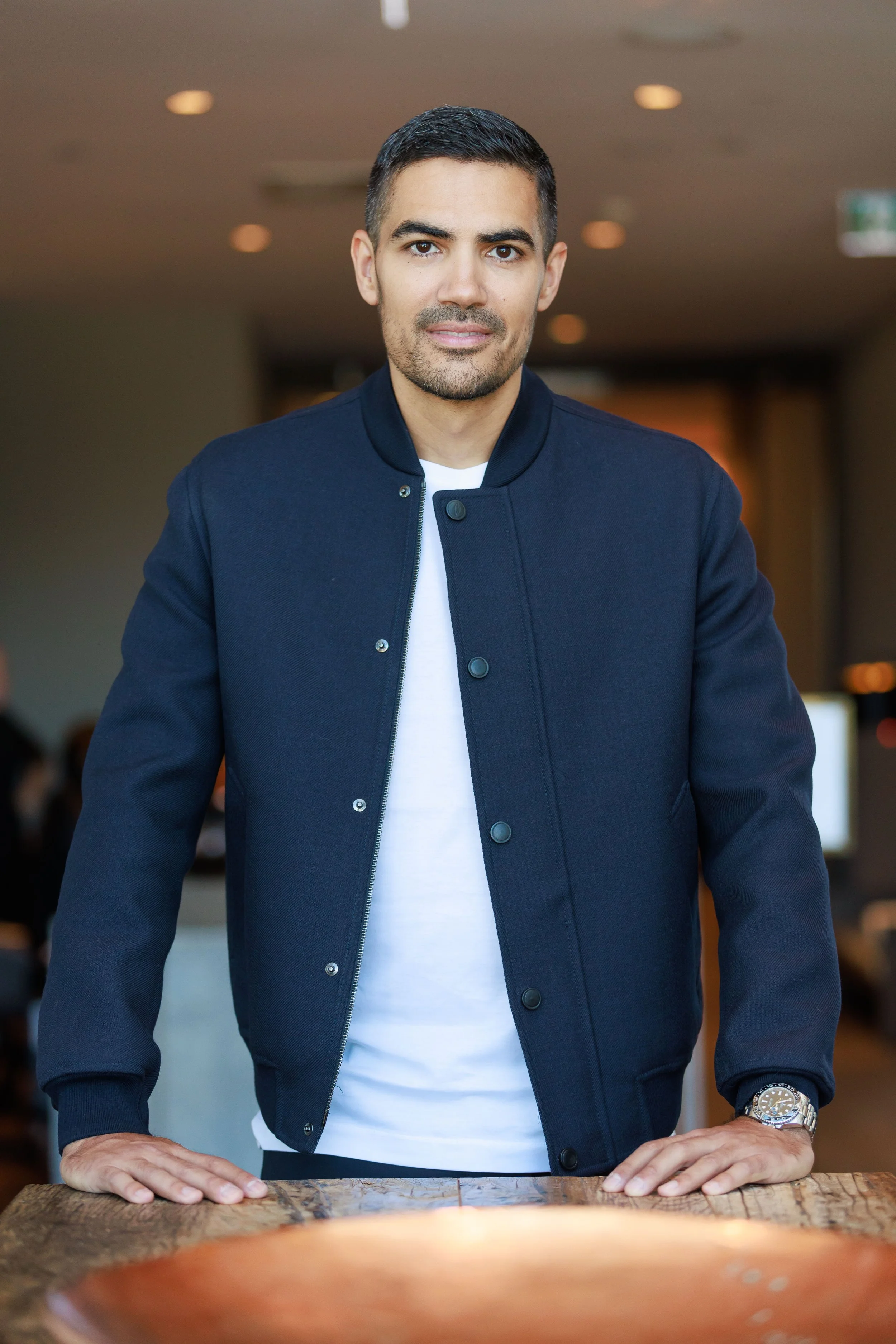 A man standing behind a wooden table, wearing a navy blue jacket, white shirt, and a watch on his left wrist, in a well-lit indoor space.