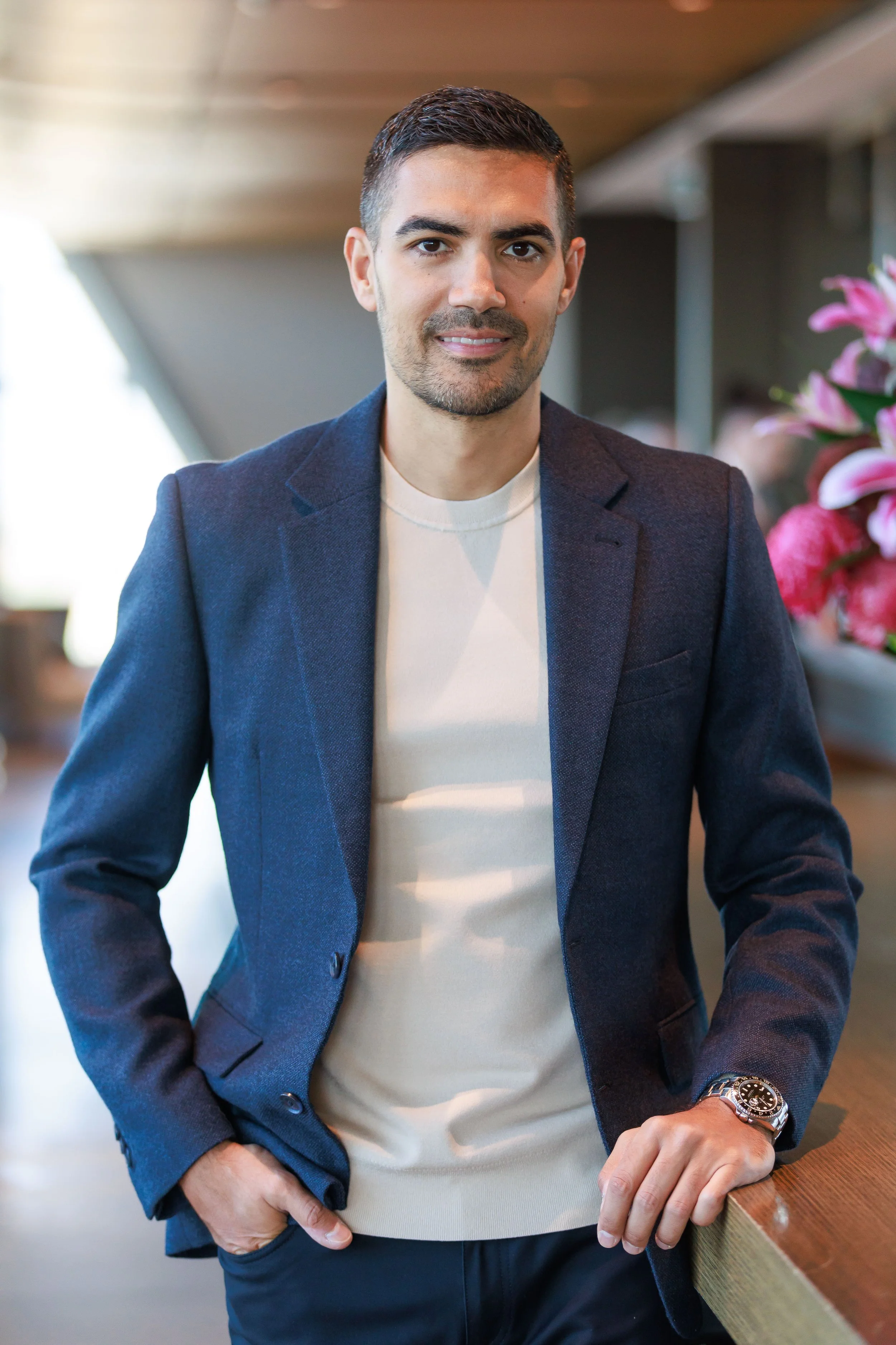 Man in a navy blazer with a watch, standing at a wooden counter with pink flowers in the background.