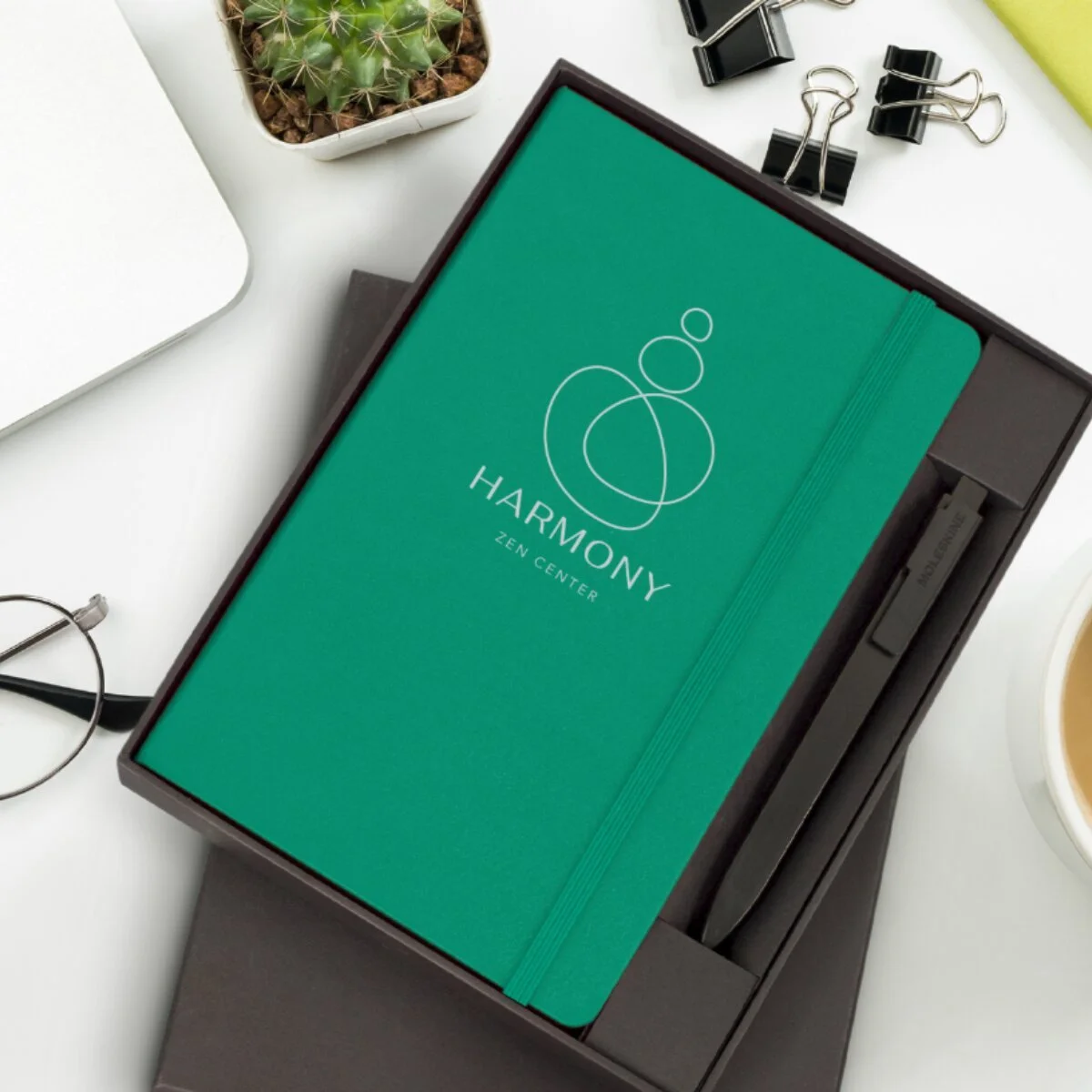A teal notebook labeled "HARMONY ZEN CENTER" is inside a black box, surrounded by office supplies including a small potted cactus, a pair of glasses, black binder clips, a yellow-green item, and a white coffee cup.