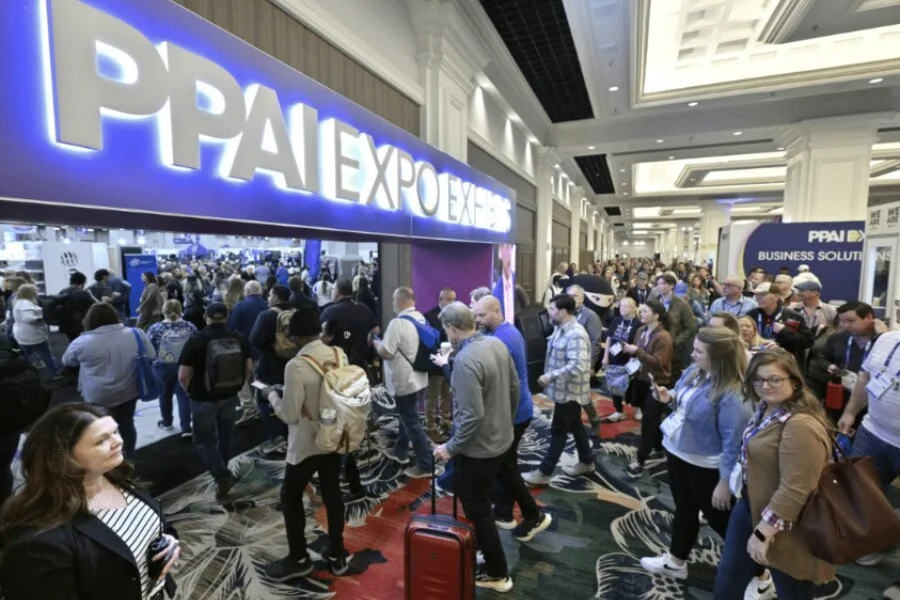 Crowd of people at PPAI Expo trade show in a hotel or conference center hallway, with illuminated signage for PPAI Expo and business solutions.
