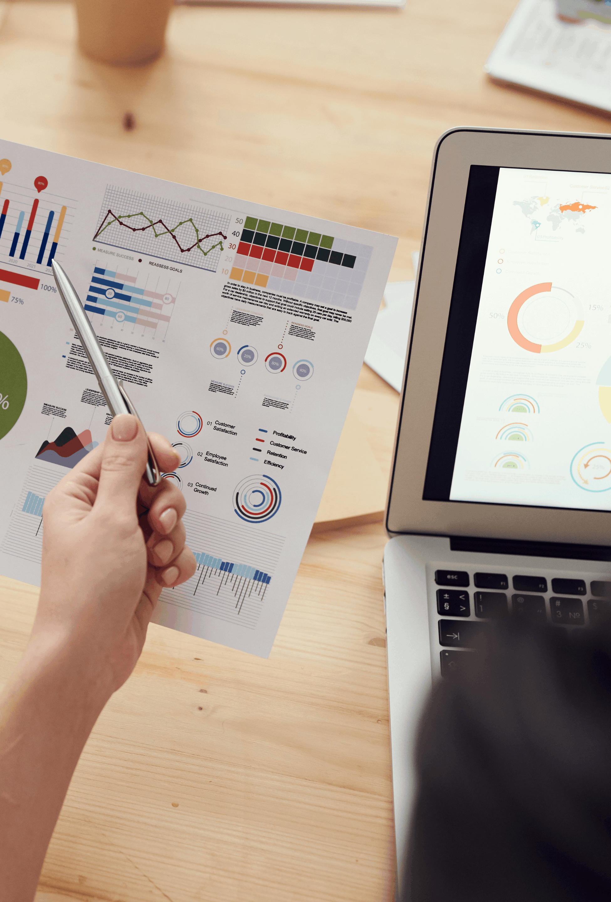 Person holding a printed sheet with colorful charts and data visuals, with a laptop displaying similar graphs on the right, on a wooden desk.