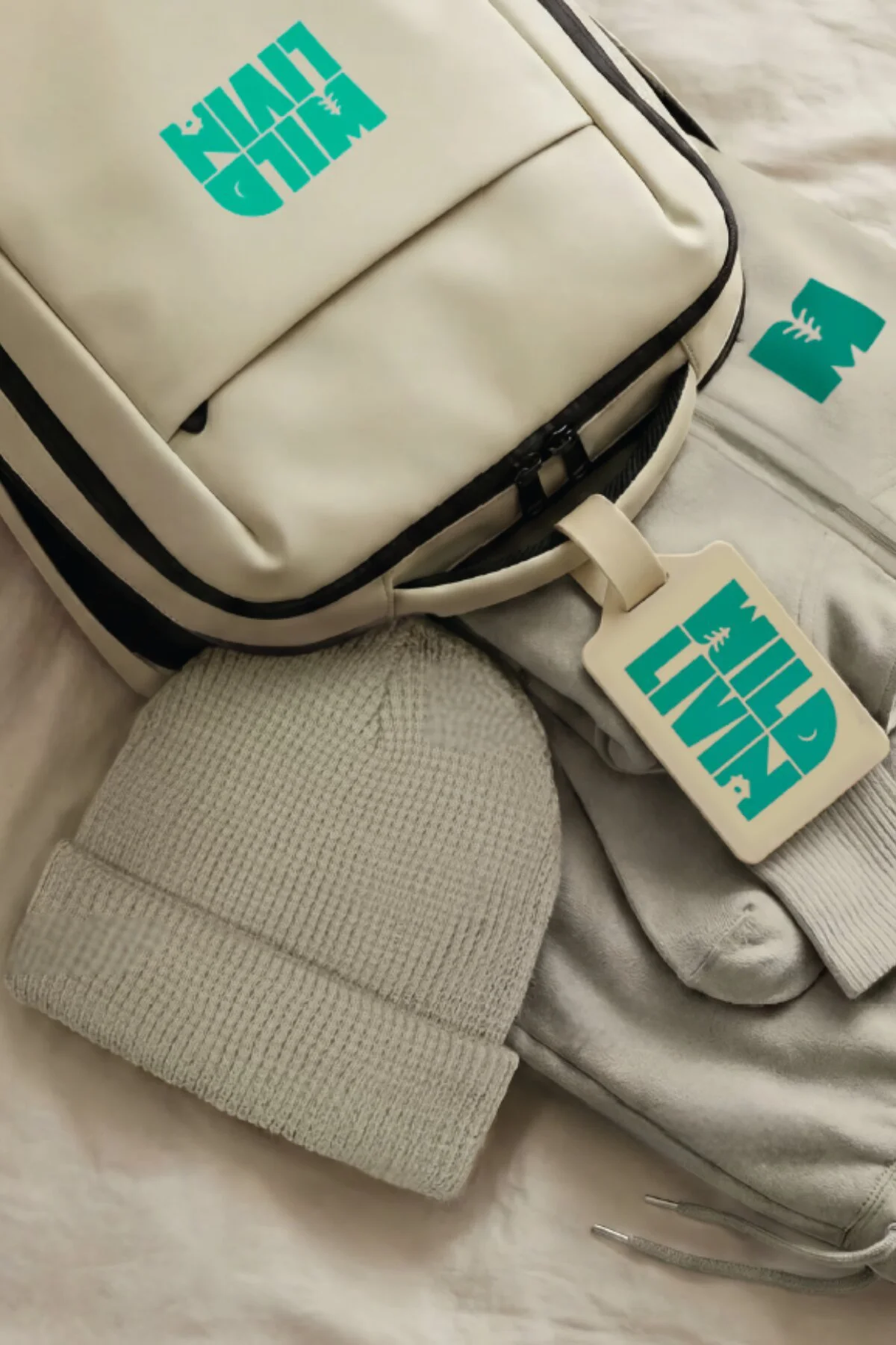 A beige backpack with light blue 'WILD CHILD' text and logo, lying on light-colored fabric. A beige knit beanie is partly visible in the foreground.