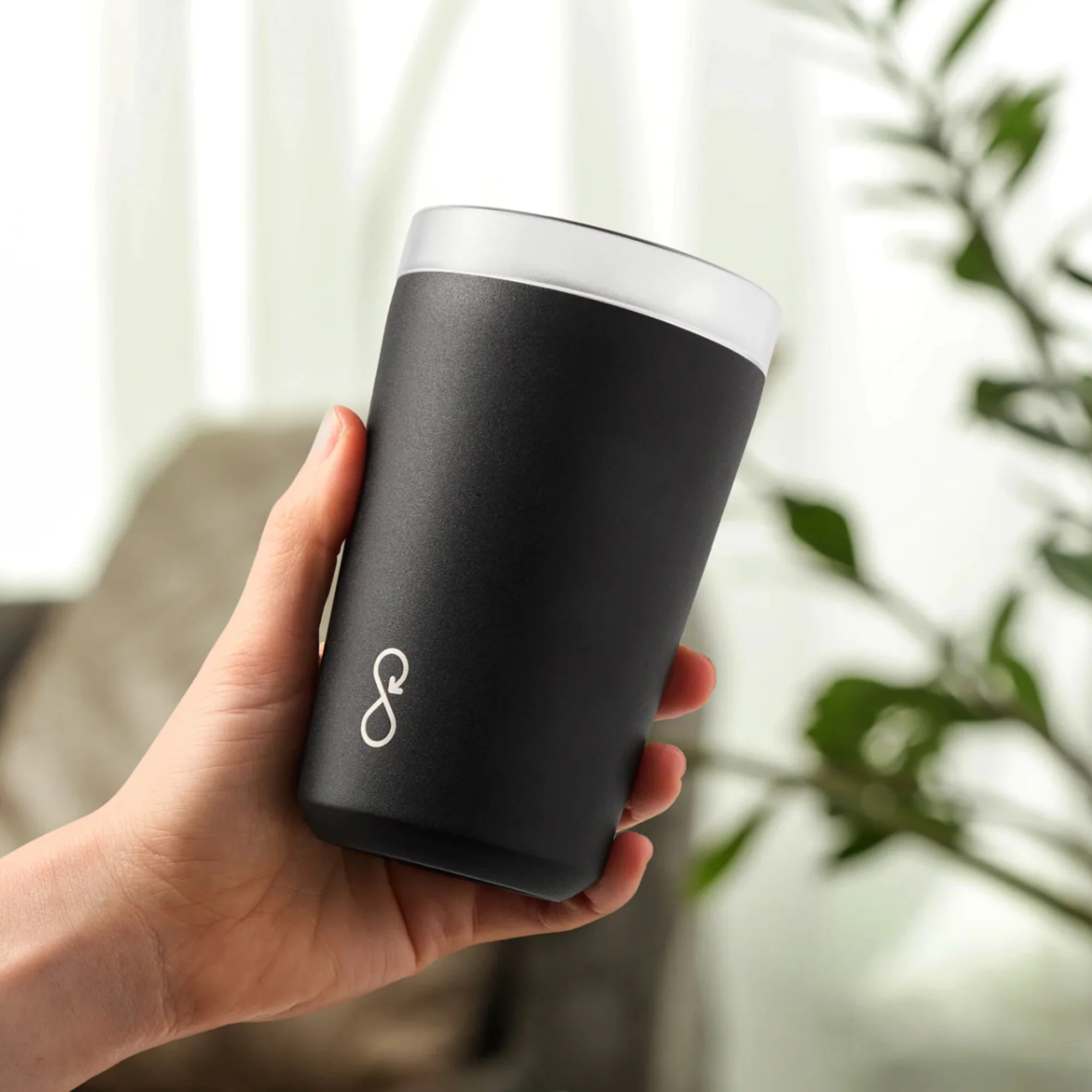 A hand holding a black tumbler with a white lid in front of a light-colored curtain and some green leaves.