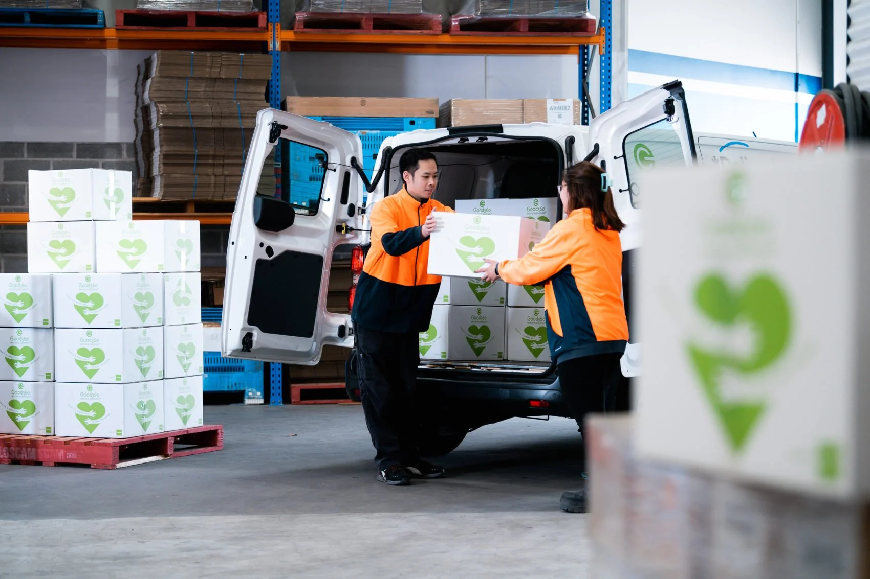 Two delivery workers in orange and black jackets loading boxes from a white van in a warehouse, surrounded by stacked cardboard boxes and pallets.