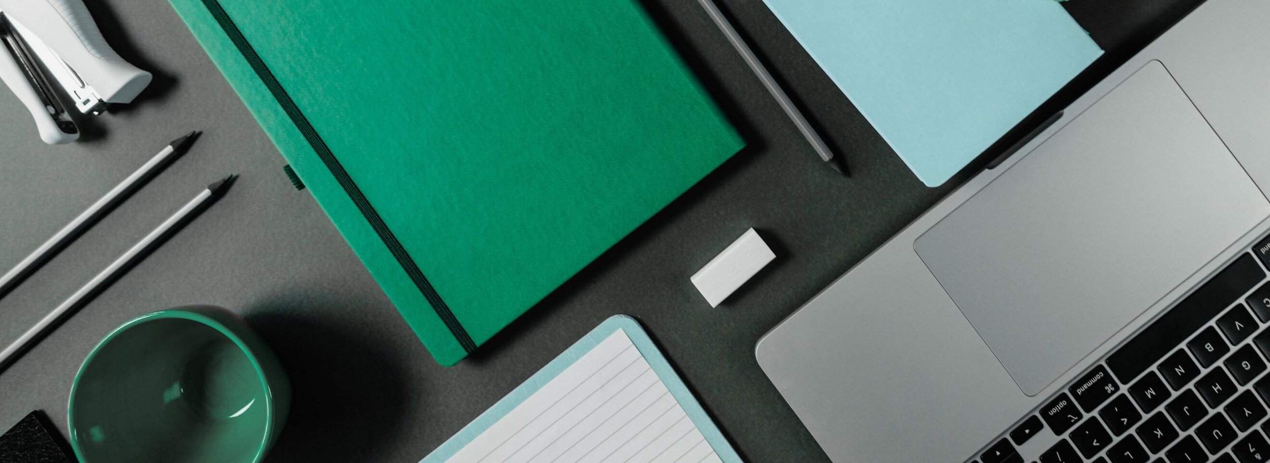 Top view of a workspace with a laptop, green notebook, black pens, white eraser, teal coffee mug, and a notepad on a grey desk.