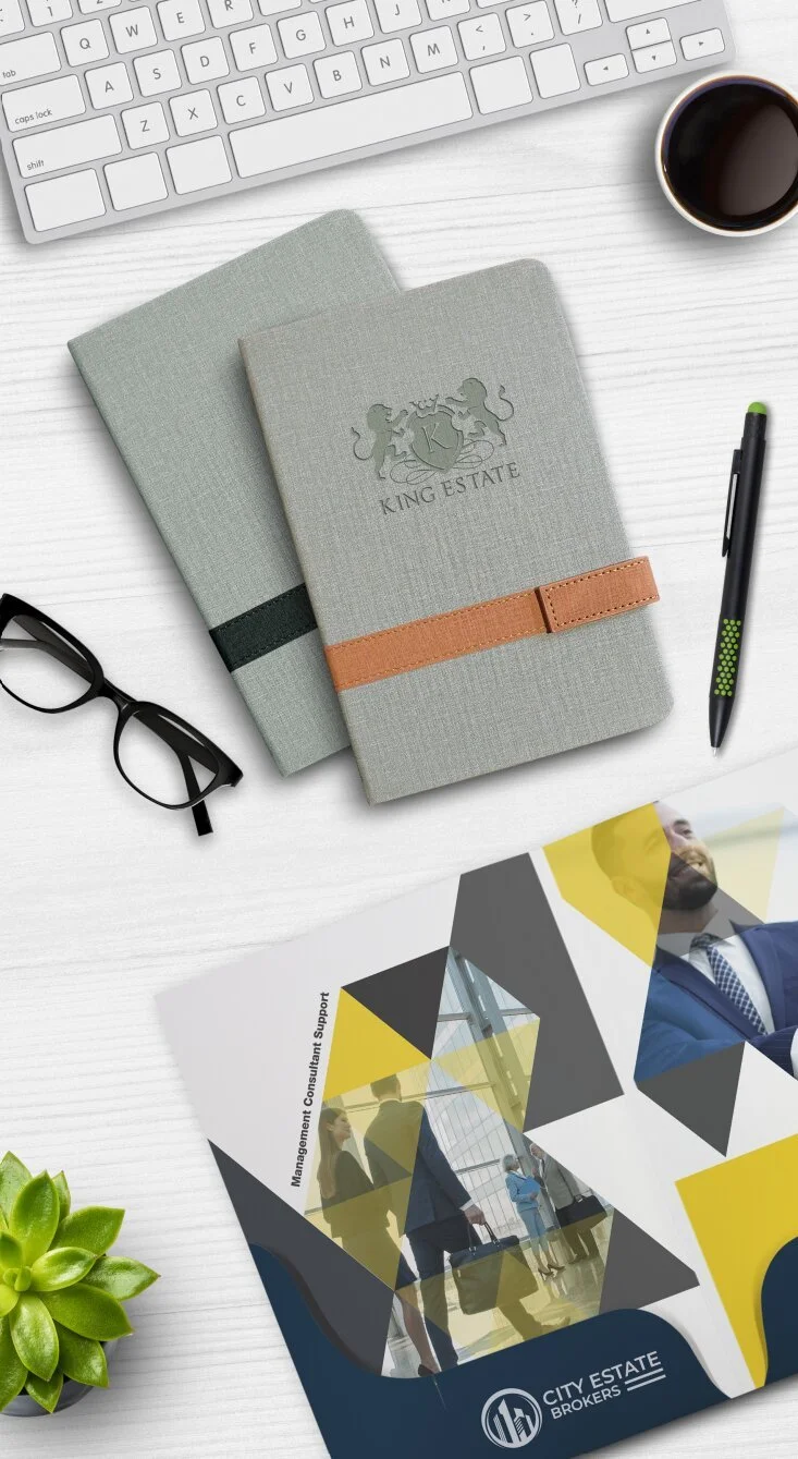 White desk with a white keyboard, a black coffee cup, two gray notebooks with a King Estate logo, black glasses, a black pen with green accents, a colorful brochure featuring people and the words "Management Consultant Support," and a green succulent plant.