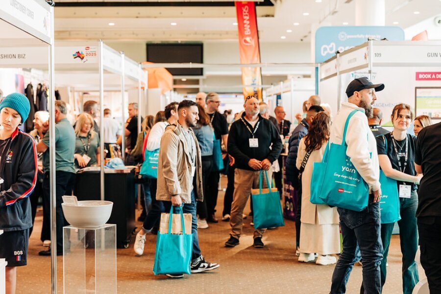 People attending a trade show or conference in an indoor exhibition hall, with booths and displays visible in the background.