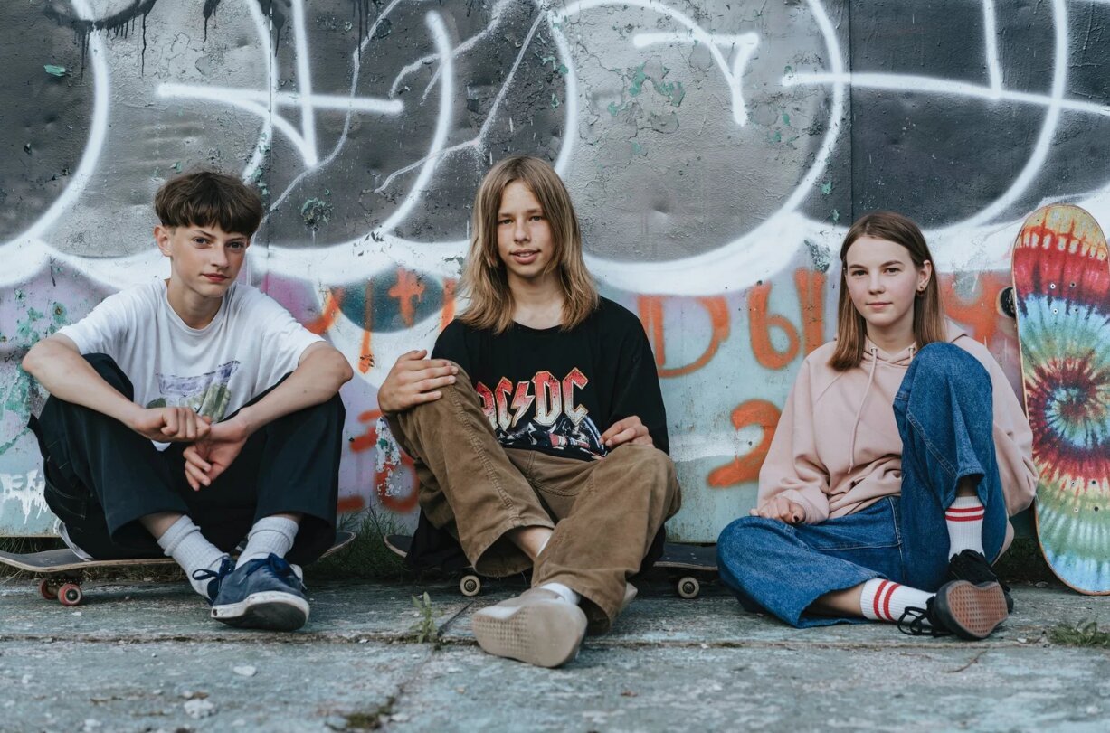 Three teenagers sitting in front of a graffiti-covered wall, with skateboards nearby. The girl on the right is holding a colorful tie-dye skateboard.