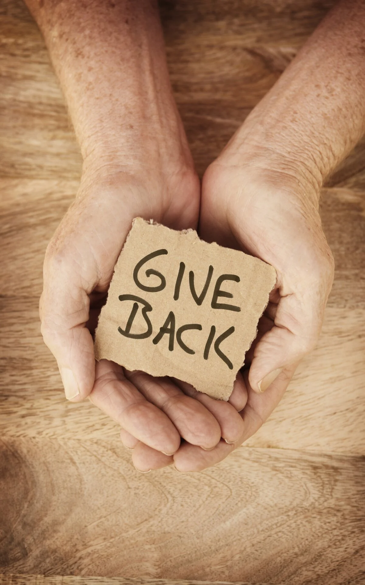 An elderly person's hands holding a torn piece of paper with the words 'GIVE BACK' handwritten on it, against a wooden background.