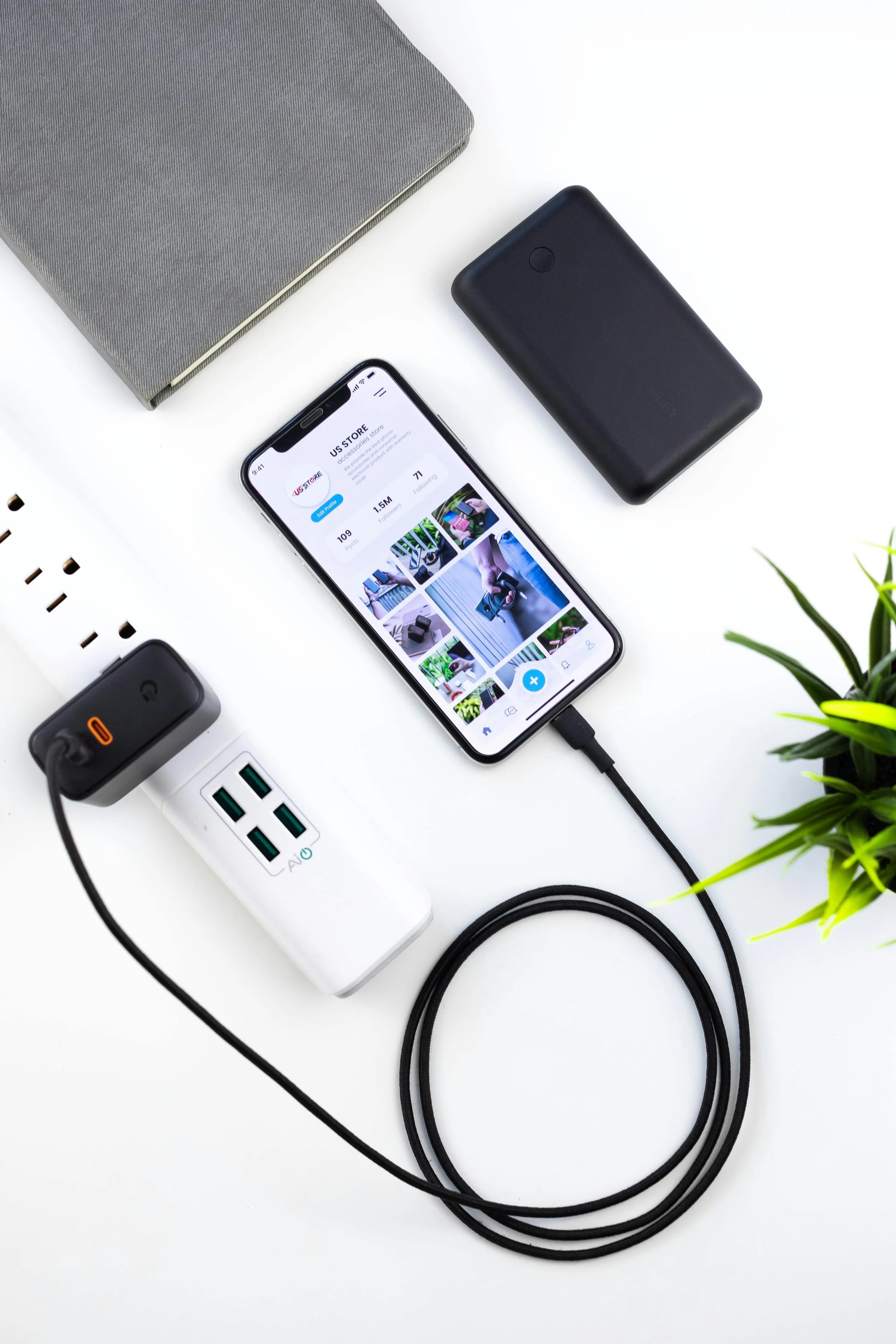 A white table with a gray laptop, an iPhone displaying a social media profile, a black portable power bank, a white power strip with multiple outlets and charging ports, and some green plant leaves in the corner.