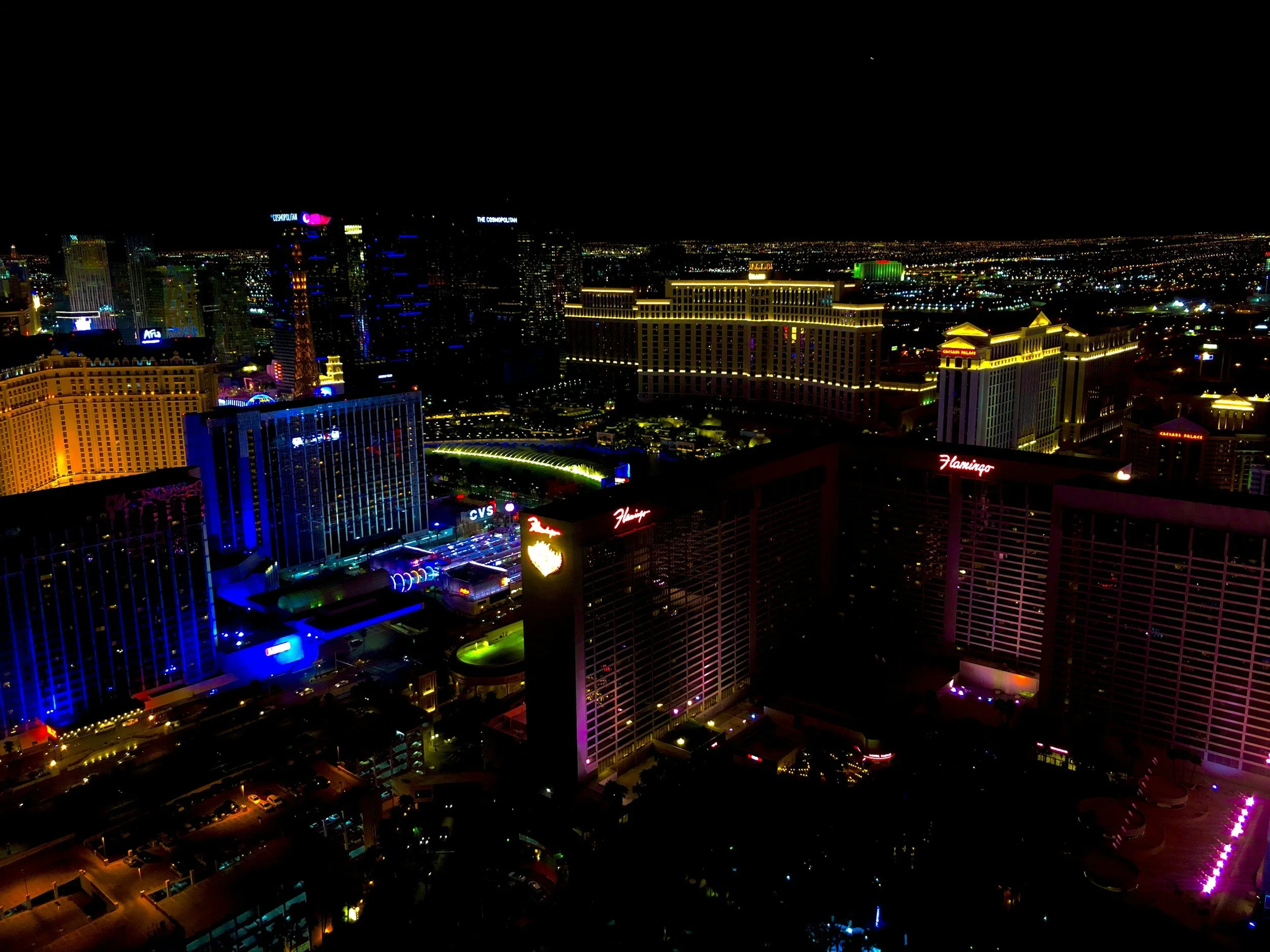 Nighttime view of Las Vegas skyline with brightly lit hotels and casinos, including Flamingo Hotel and CVS, under a dark sky with city lights in the distance.