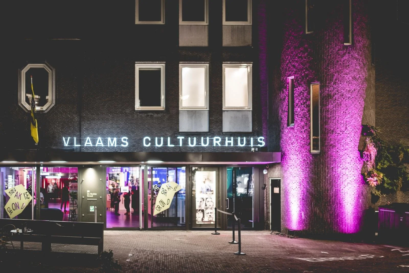 Vlaams Cultuurhuis entrance at night with illuminated sign and pink-purple lighting on the building, displaying a lively interior scene with people gathered inside.