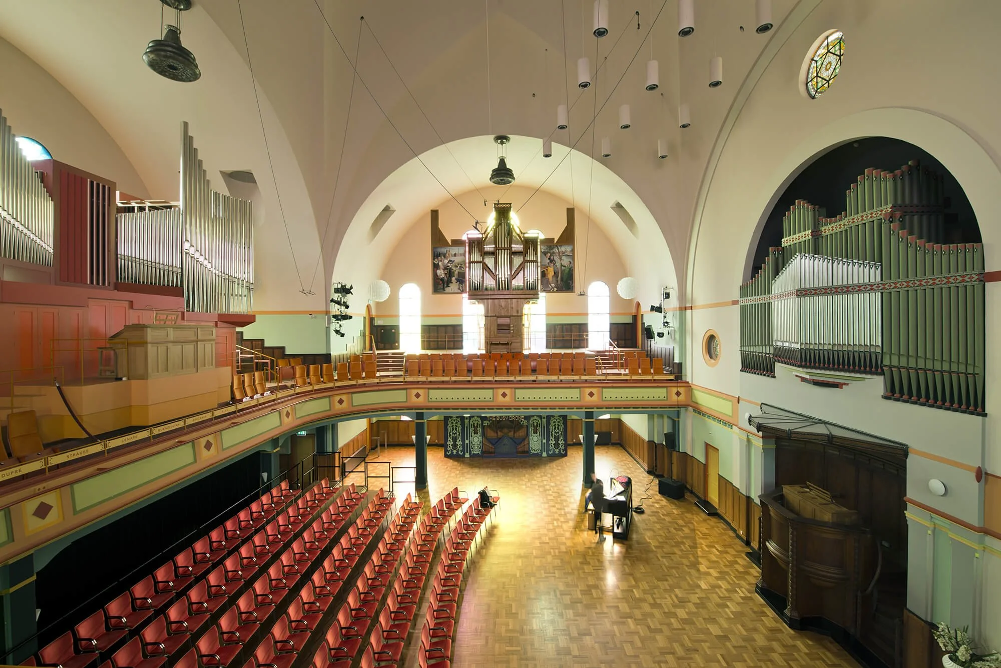 Interior of a concert hall with a large pipe organ, wooden flooring, and red chairs. A musician is playing the piano on stage. The hall features arched ceilings and stained glass windows.