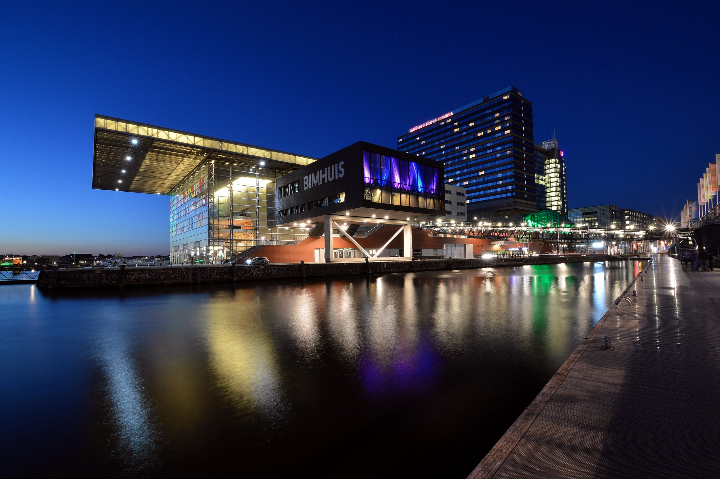 Bimhuis concert venue in Amsterdam at night, reflecting on the water, with surrounding buildings and a clear blue sky.