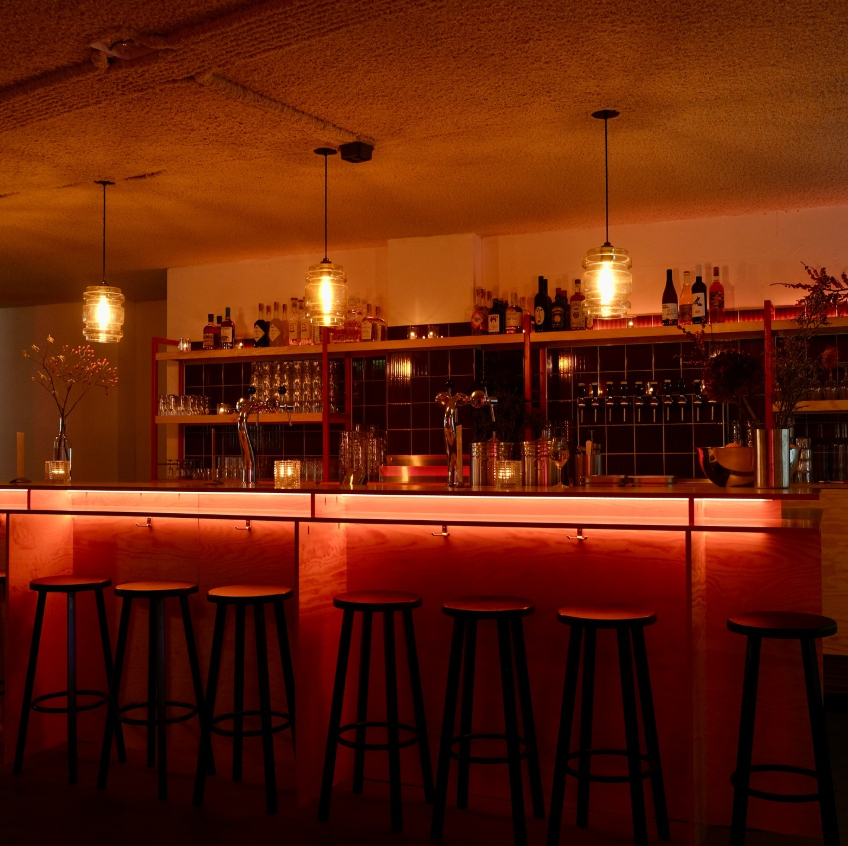 Cozy bar interior with stools, dim lighting, hanging lamps, and bottles on shelves.