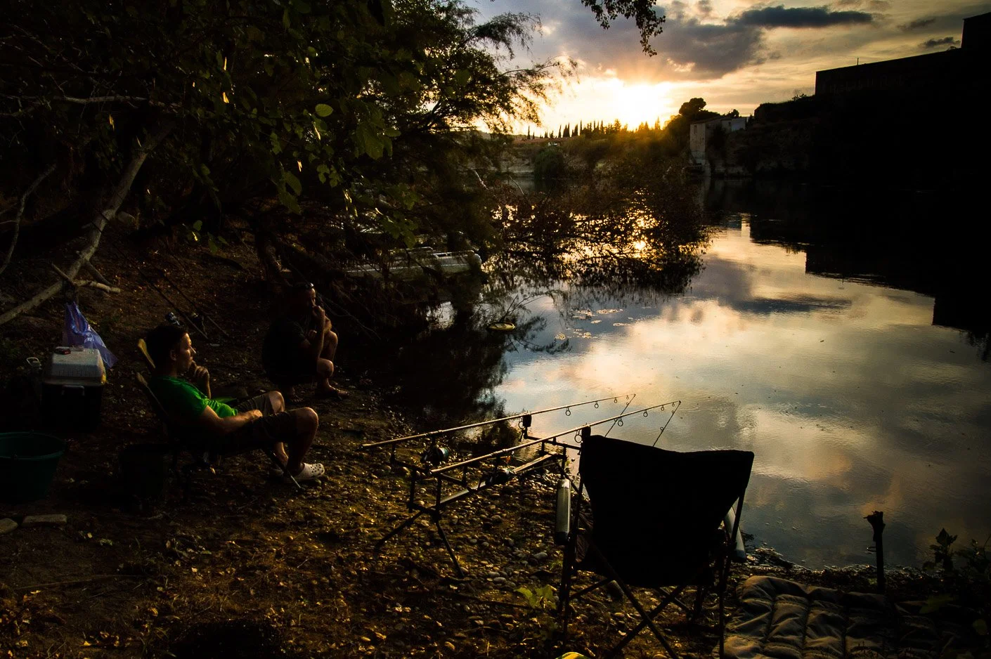 Two people fishing by a river at sunset, with fishing rods set up on the shore and a chair nearby, under trees with a reflective water surface and a cloudy sky.