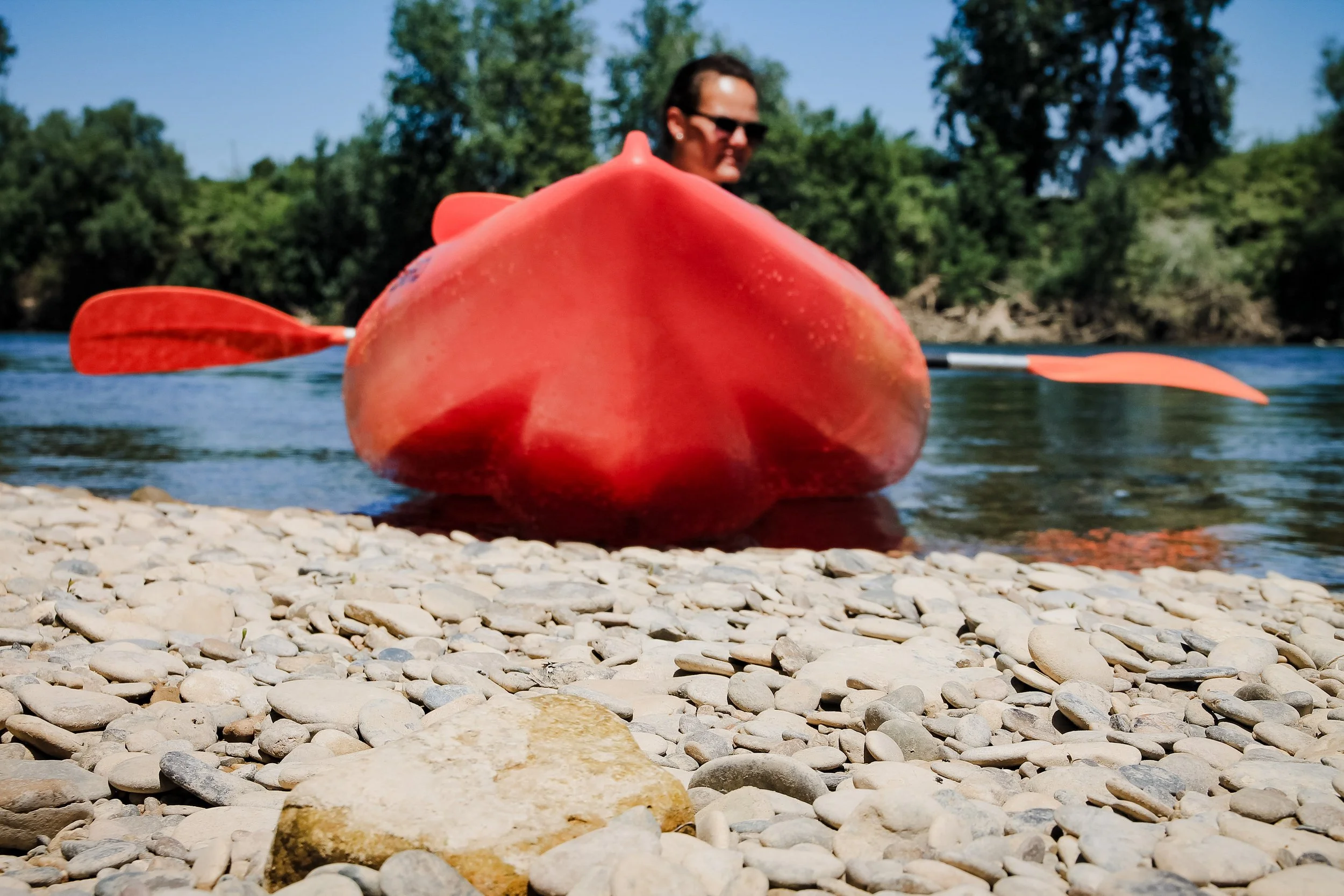 A man with sunglasses sitting in a red kayak on a river with a pebble beach in the foreground and trees in the background.